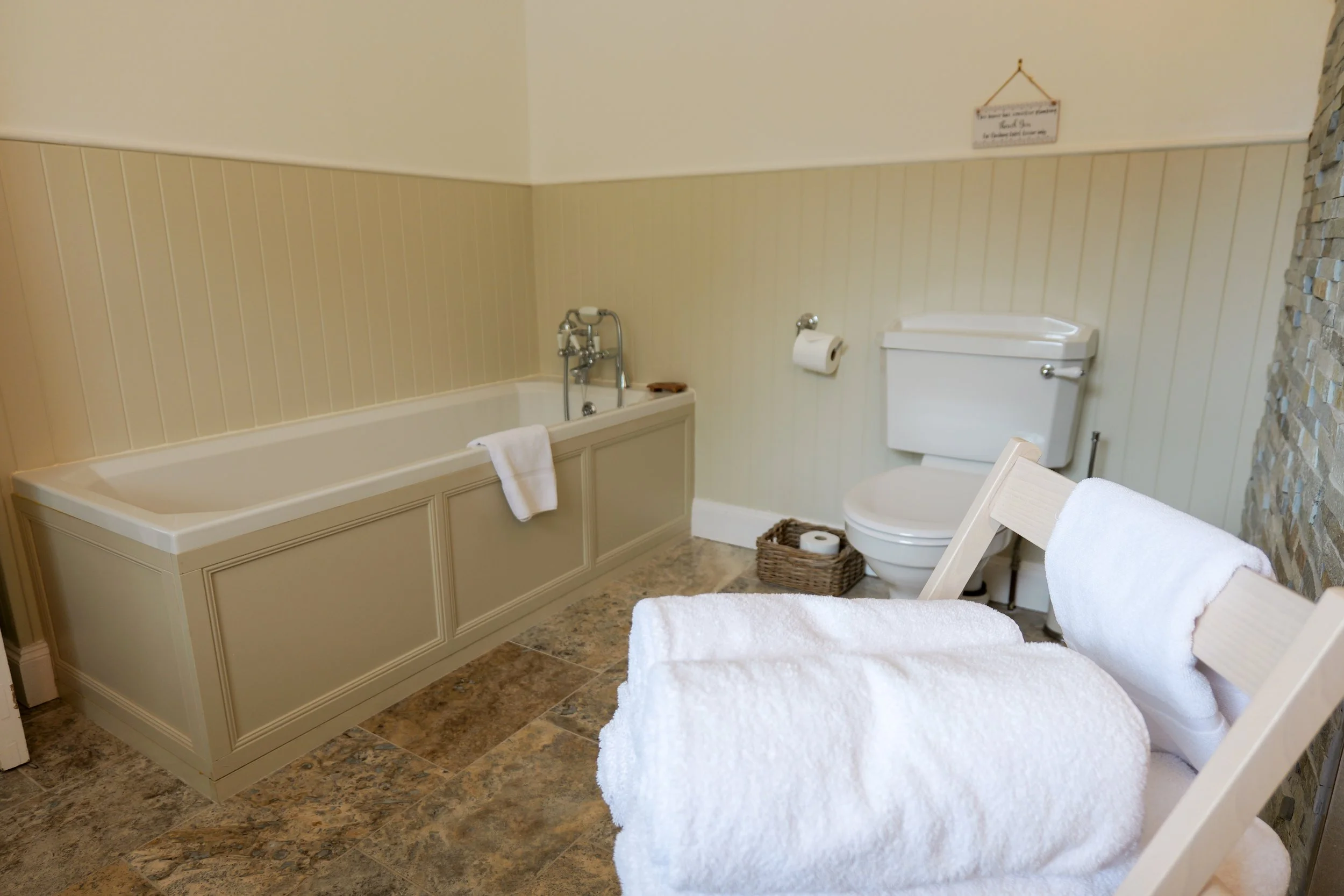 A bathroom with a bathtub, toilet, and white towels on chairs, with a stone accent wall and beige-paneled walls.