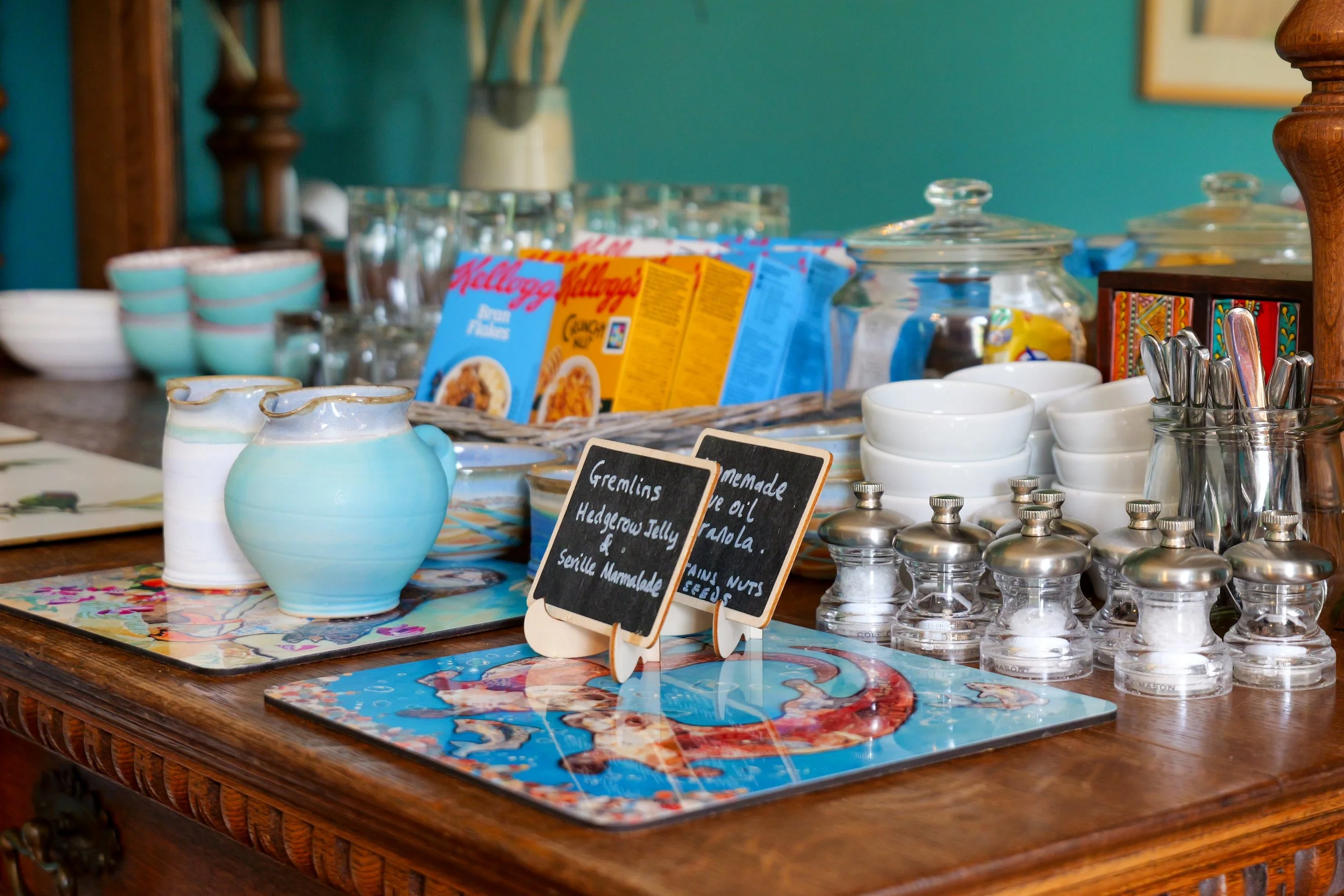 A table set with various dishes, bowls, and utensils. Two small chalkboards display handwritten labels of jelly and homemade oil and banana. Behind are cereal boxes and glass jars.