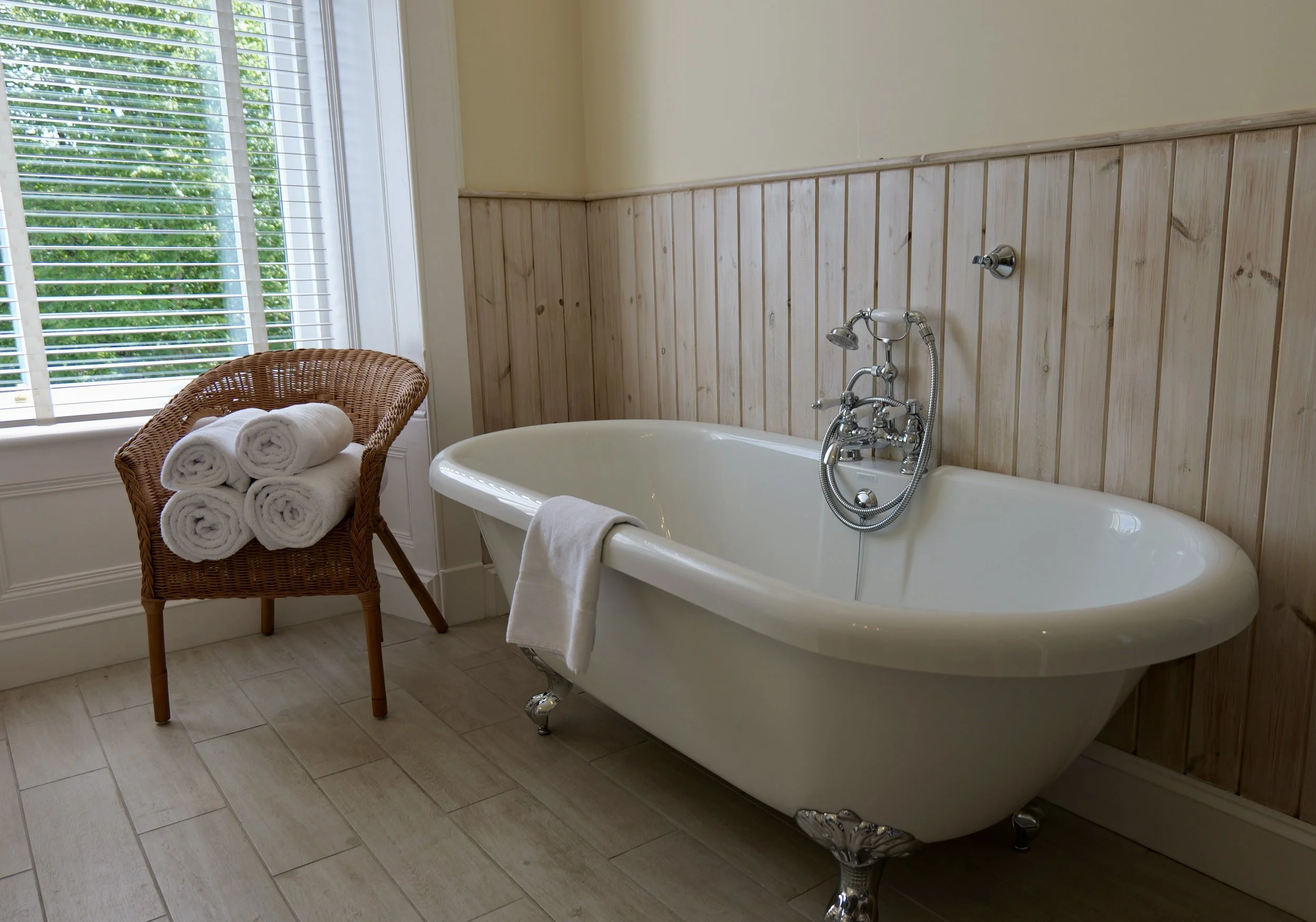 Bathroom with a white clawfoot bathtub, a wicker chair holding rolled white towels, a window with blinds, and wooden wall paneling.