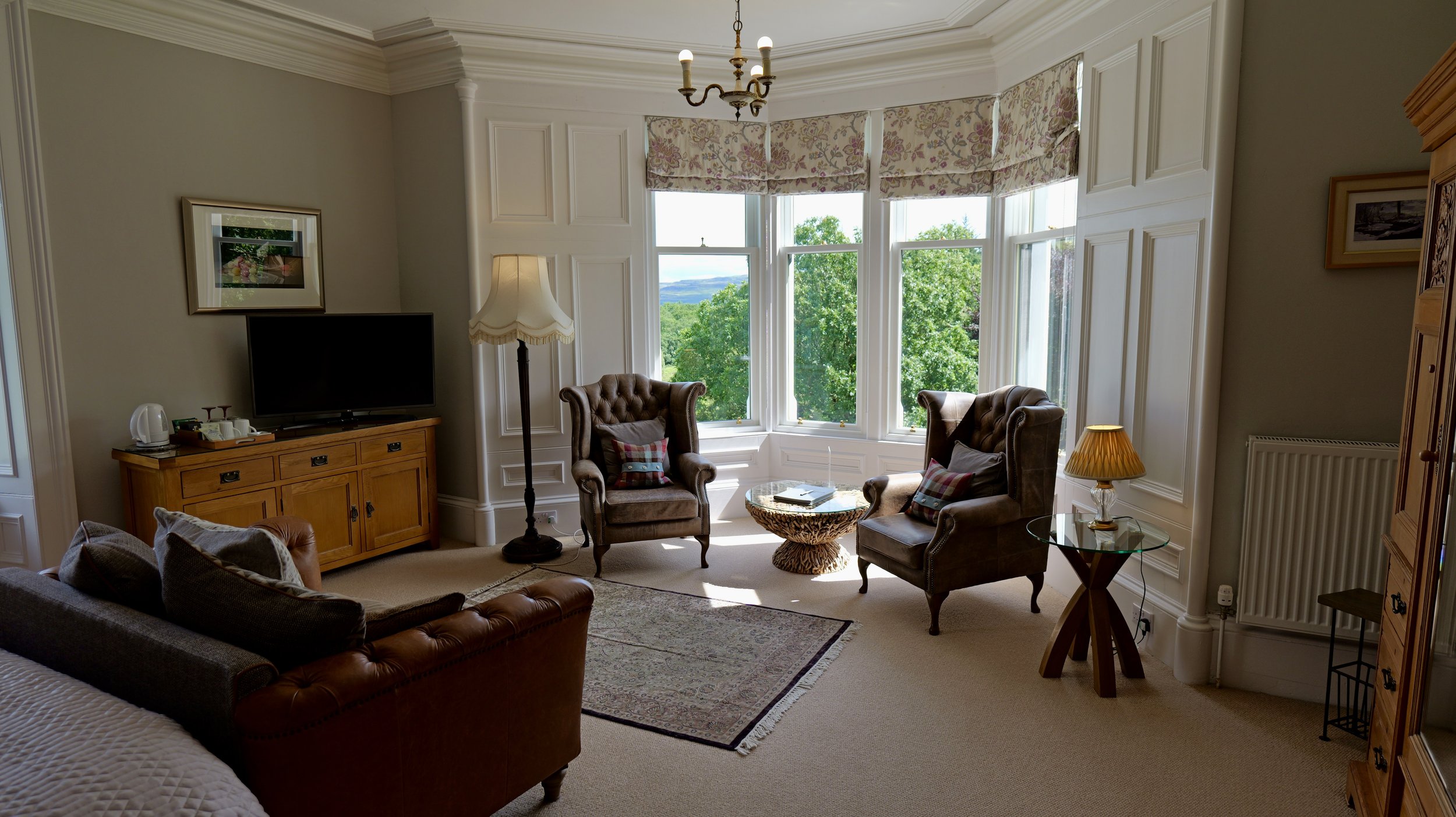 Living room with bay window, two brown armchairs, a sofa with pillows, a wooden TV stand with a flat-screen TV, a standing lamp, a side table with a yellow lamp, and a view of green trees and hills outside.