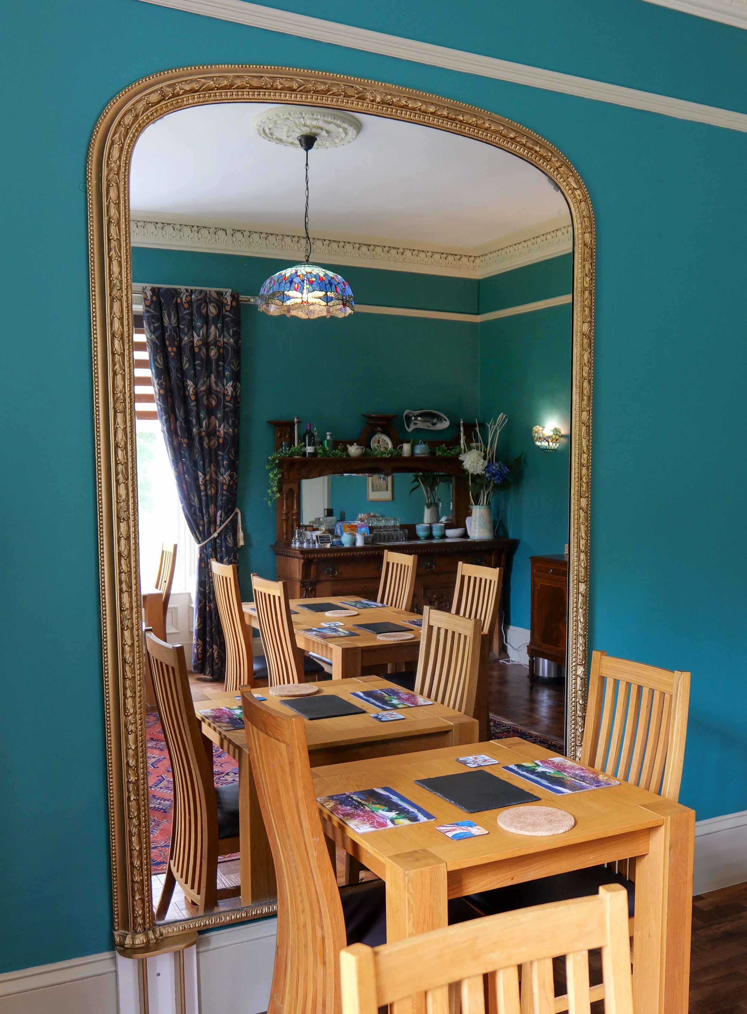 Dining room viewed through a large ornate gold-framed mirror, with a wooden dining table and six matching chairs, set with placemats and postcards, against teal walls with white crown molding.
