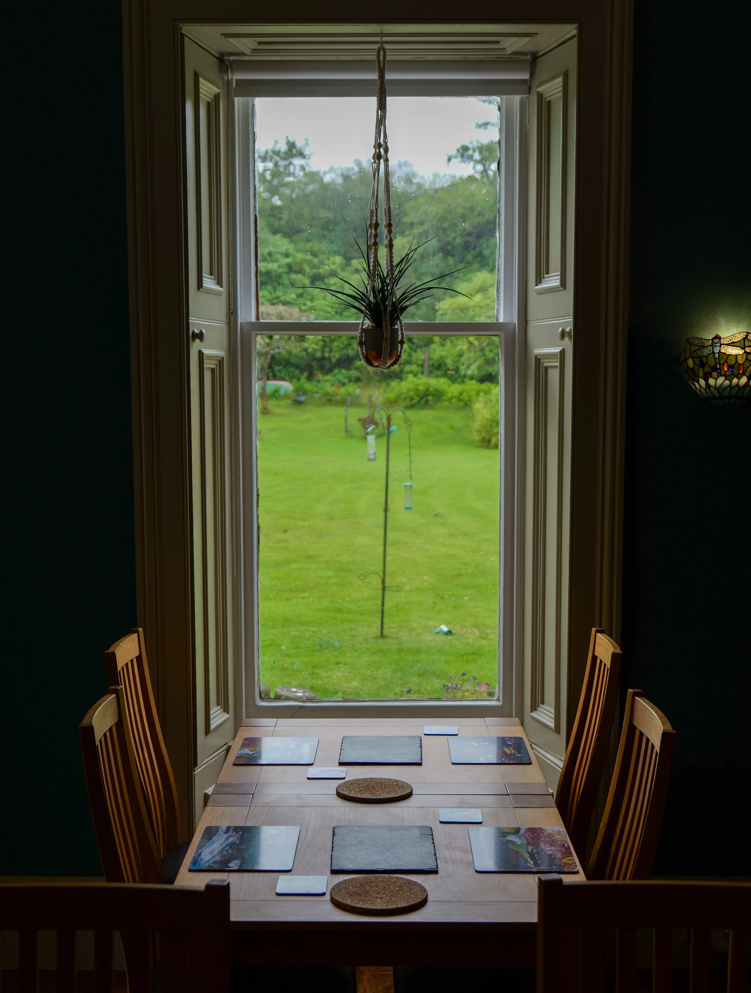 A view from inside of a dining area with a wooden table set with six placemats and two circular cork coasters. The table is in front of a window with open shutters, overlooking a green grassy yard with trees and a few bird feeders. A hanging plant is