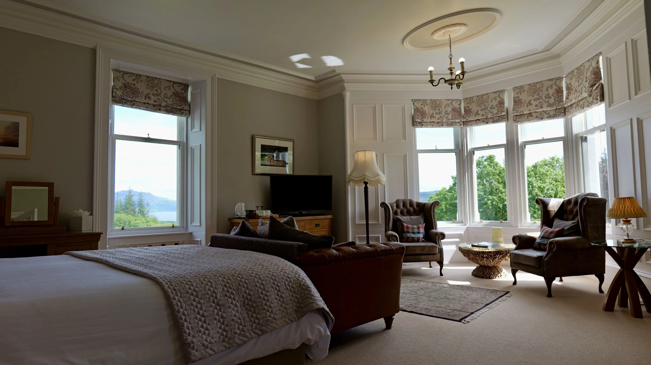 A cozy bedroom with large windows overlooking greenery and mountains, featuring a bed, armchairs, a TV, lamps, and decorative touches.