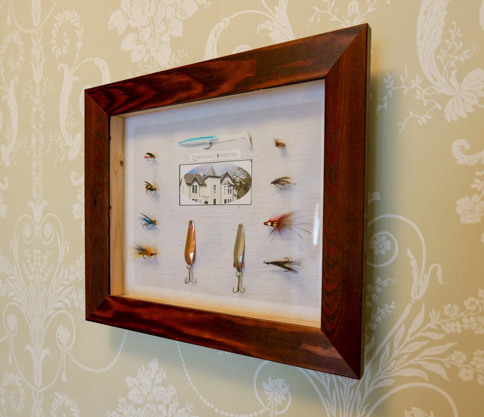 Display case with fishing lures and a photo of a house, labeled 'Lindhu House', mounted on a patterned beige wall.