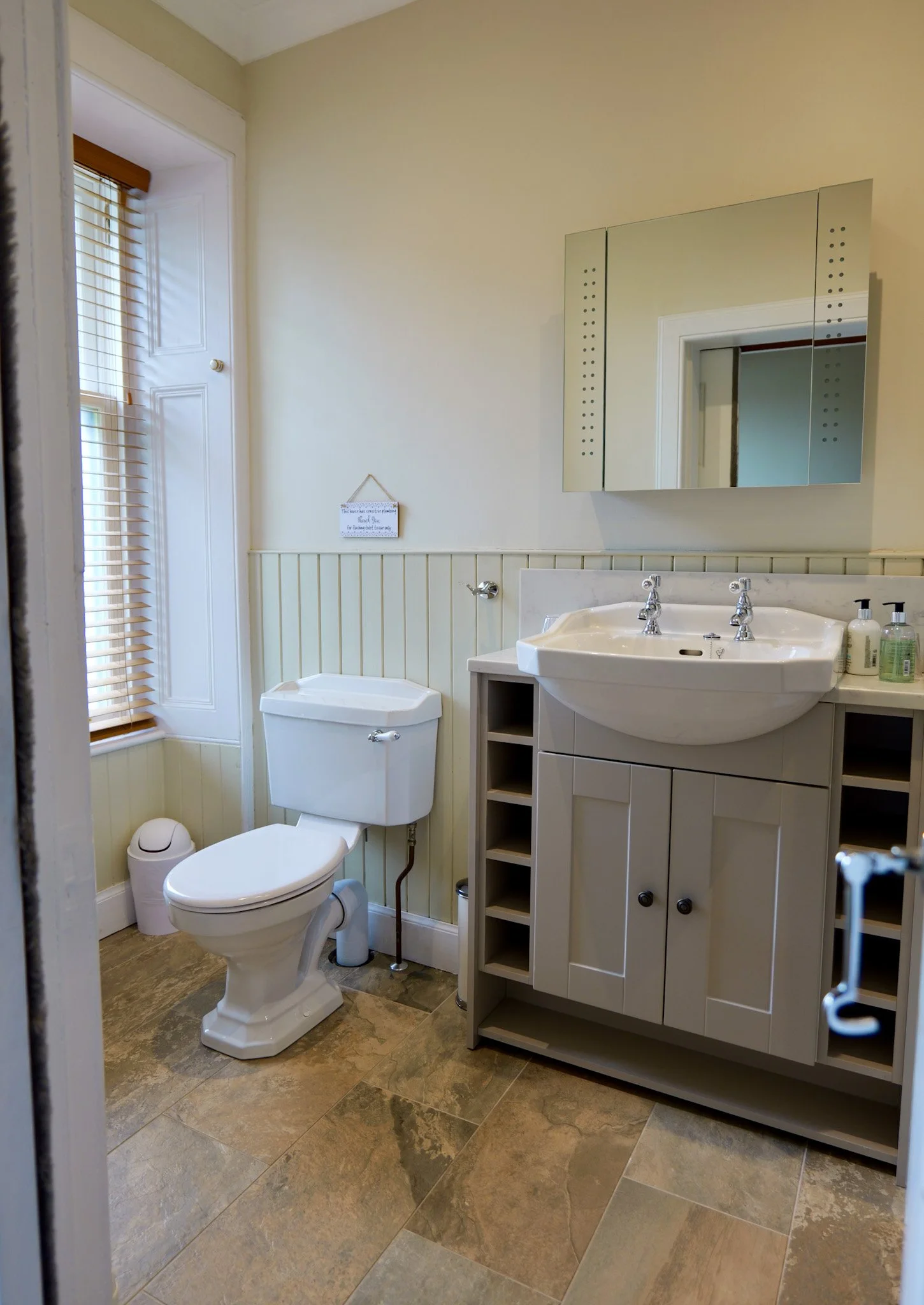 A small bathroom with a toilet, a sink with a cabinet below, a mirror, a window with blinds, and a small trash can on the floor. The walls are partially paneled with vertical beadboard, painted cream, and the flooring is stone tile.
