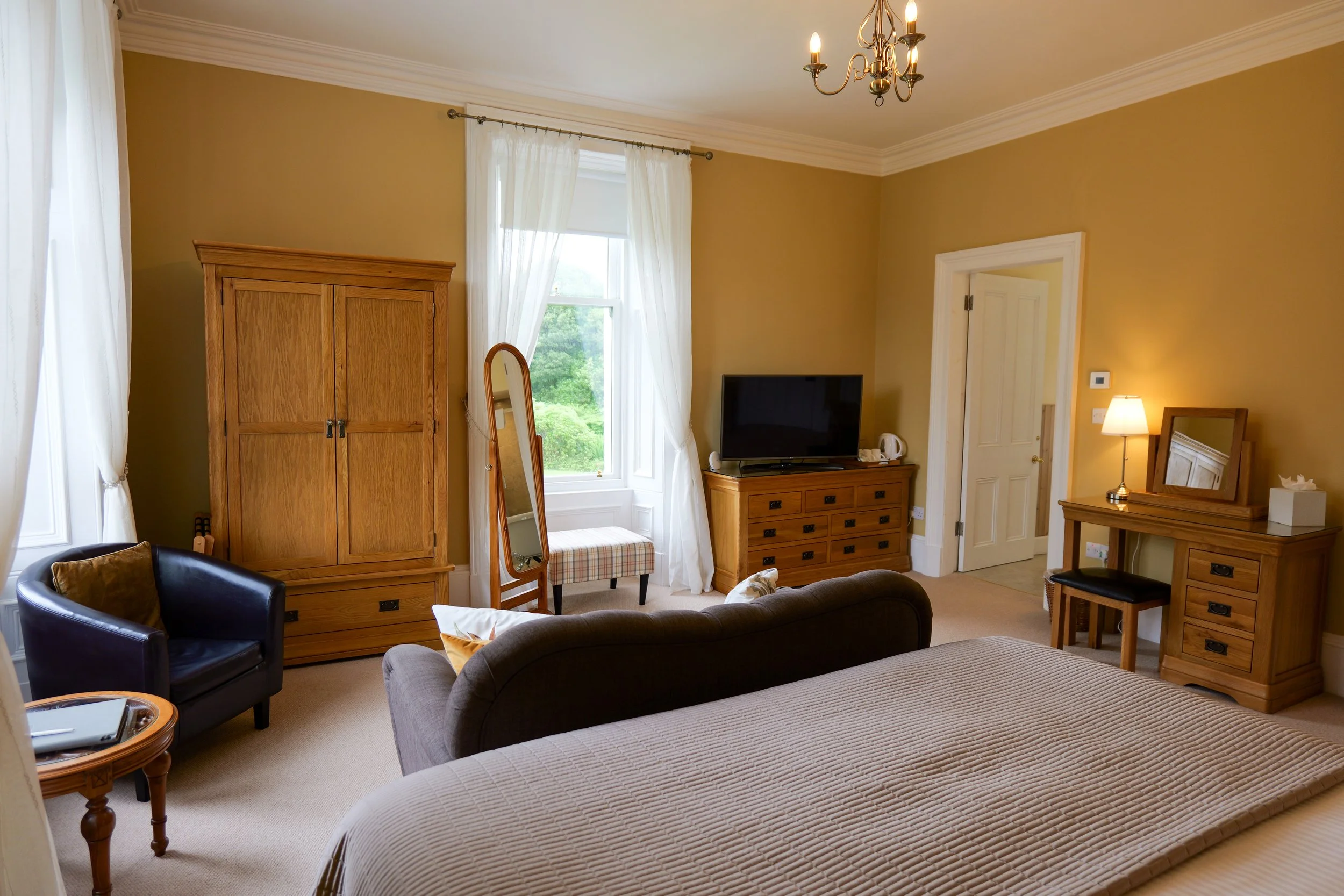 A cozy bedroom with beige walls, a bed with a beige bedspread, a wooden wardrobe, a full-length mirror, a window with white curtains, a TV on a wooden dresser, a small desk with a lamp and a mirror, and a black armchair.