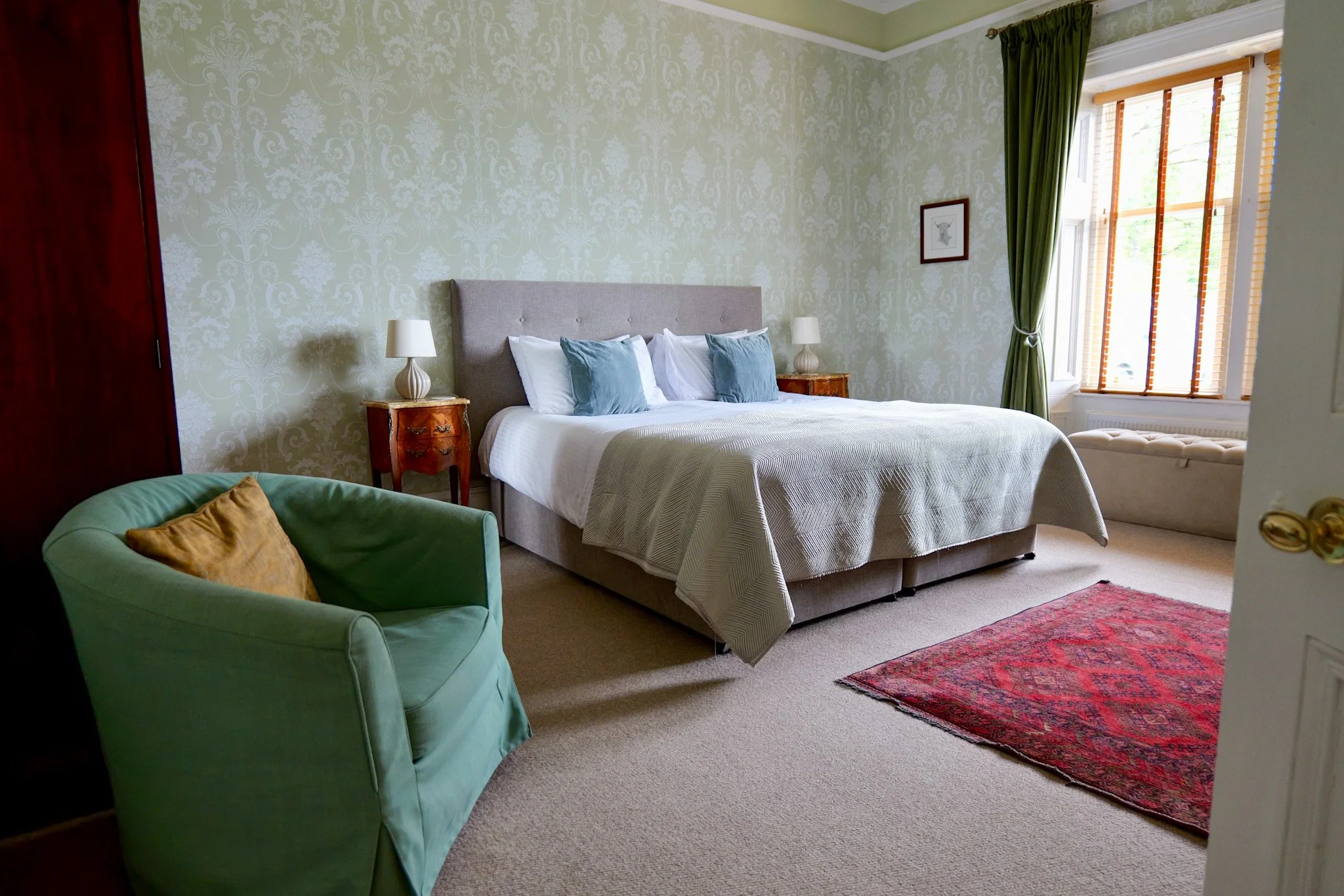 A cozy bedroom with a large bed featuring a gray upholstered headboard, white bedding, and two blue pillows. Beside the bed are wooden nightstands with white lamps. Green curtains frame a window with wooden blinds, allowing natural light. There is a 