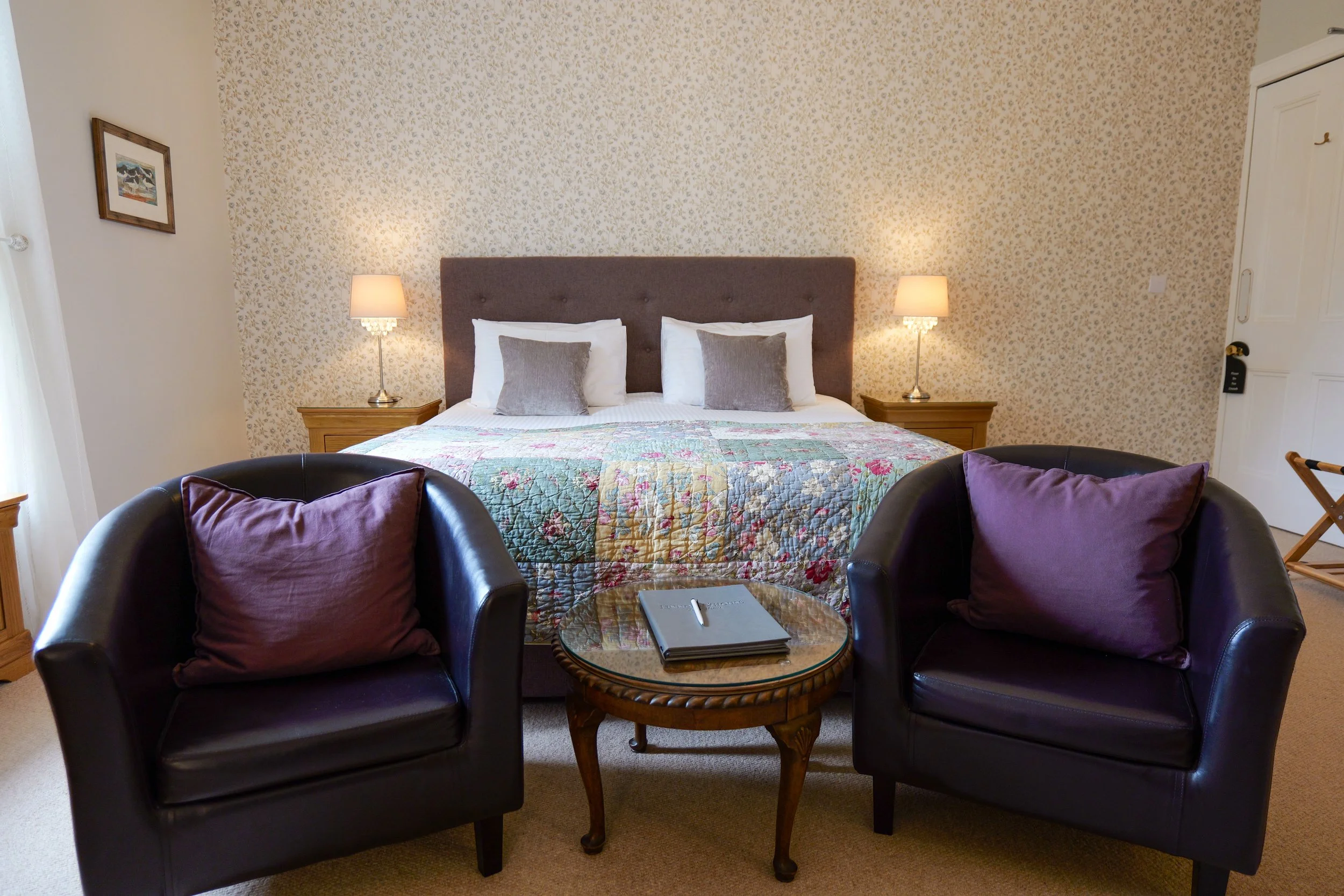 A cozy hotel room with a bed, two nightstands with lamps, two leather chairs with purple pillows, and a small glass-topped table, beige carpet, patterned wallpaper, and a wooden picture frame on the wall.