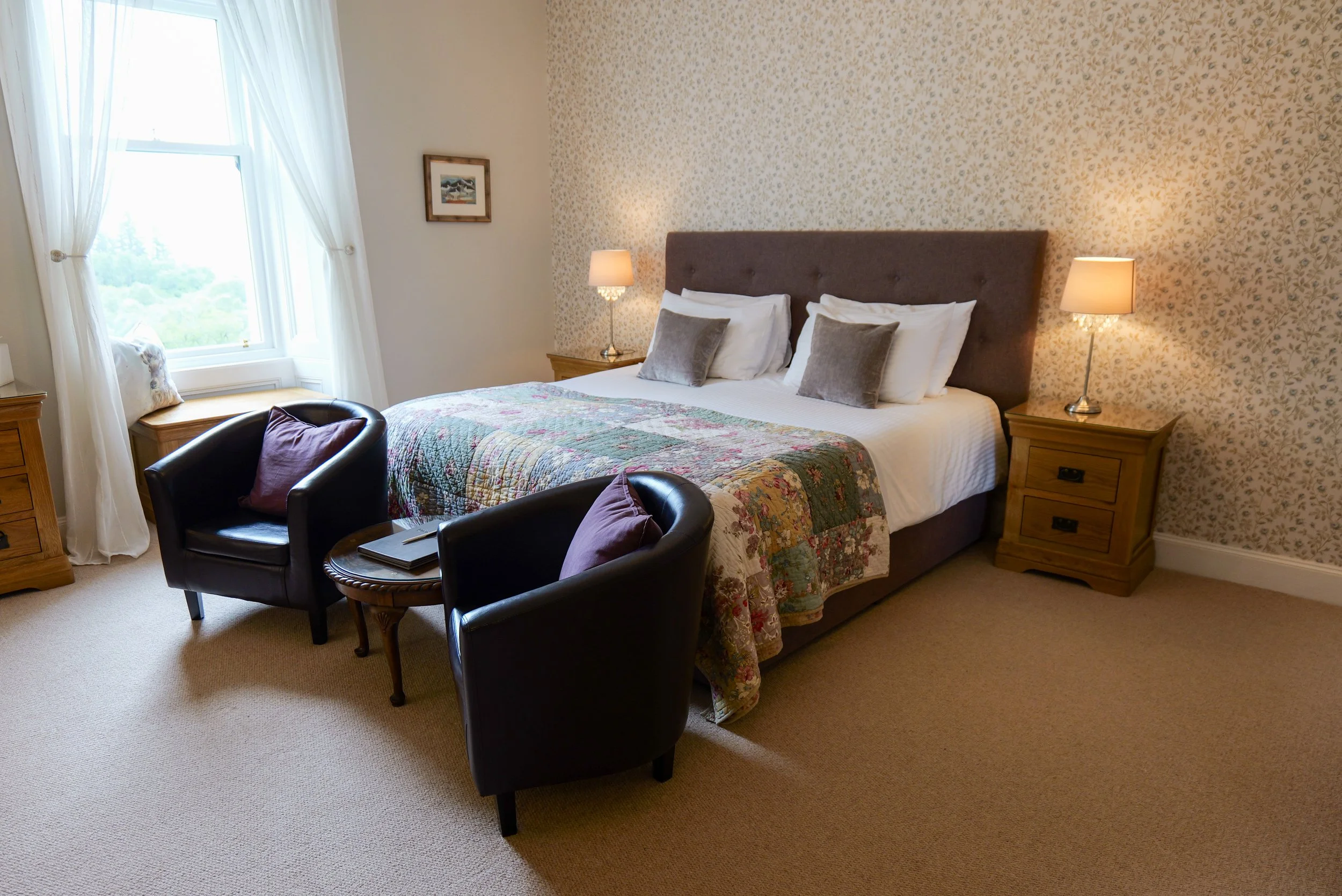 A cozy bedroom with a large bed, floral quilt, and four pillows, flanked by wooden nightstands with lamps. Two black armchairs with purple pillows sit in front of a window with white curtains, and a small table with books is between them. The wall be