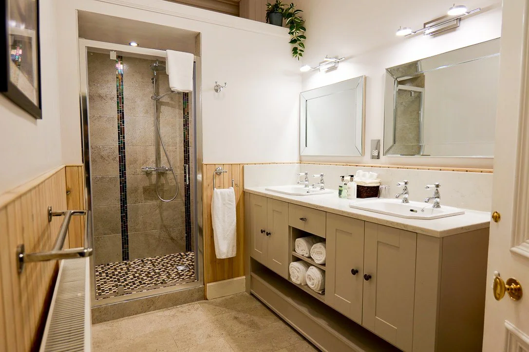 A clean, modern bathroom with a double vanity, large mirrors, and a walk-in shower with tiled walls and a small mosaic floor.