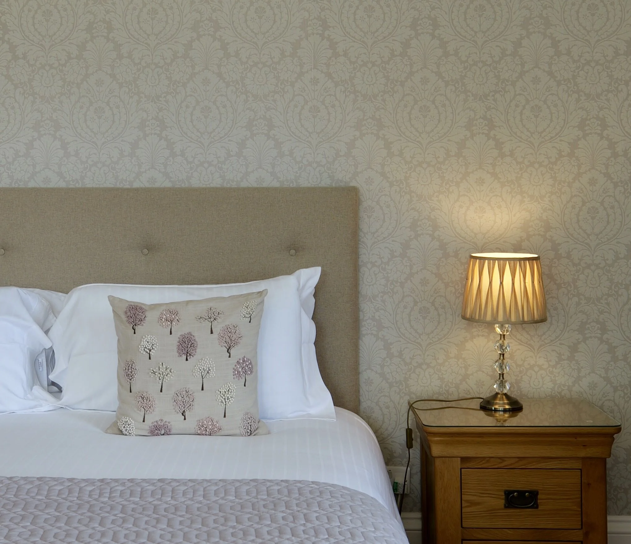 A bed with white pillows and a decorative pillow with tree patterns, beside a wooden nightstand with a lit table lamp, in a room with beige patterned wallpaper.