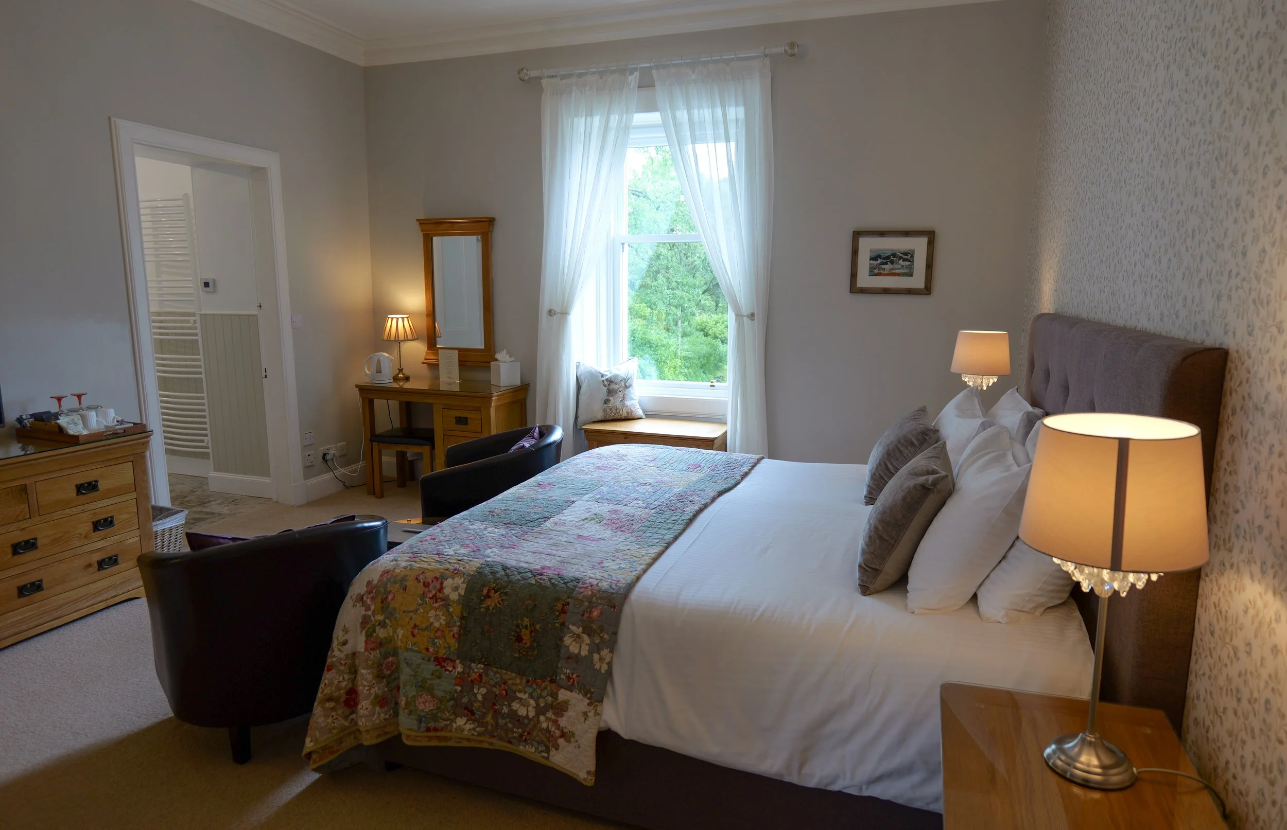 A cozy bedroom with a bed featuring white bedding and decorative pillows, a window with sheer curtains showing greenery outside, wooden furniture including bedside tables and a dresser, two black armchairs, and warm lighting.