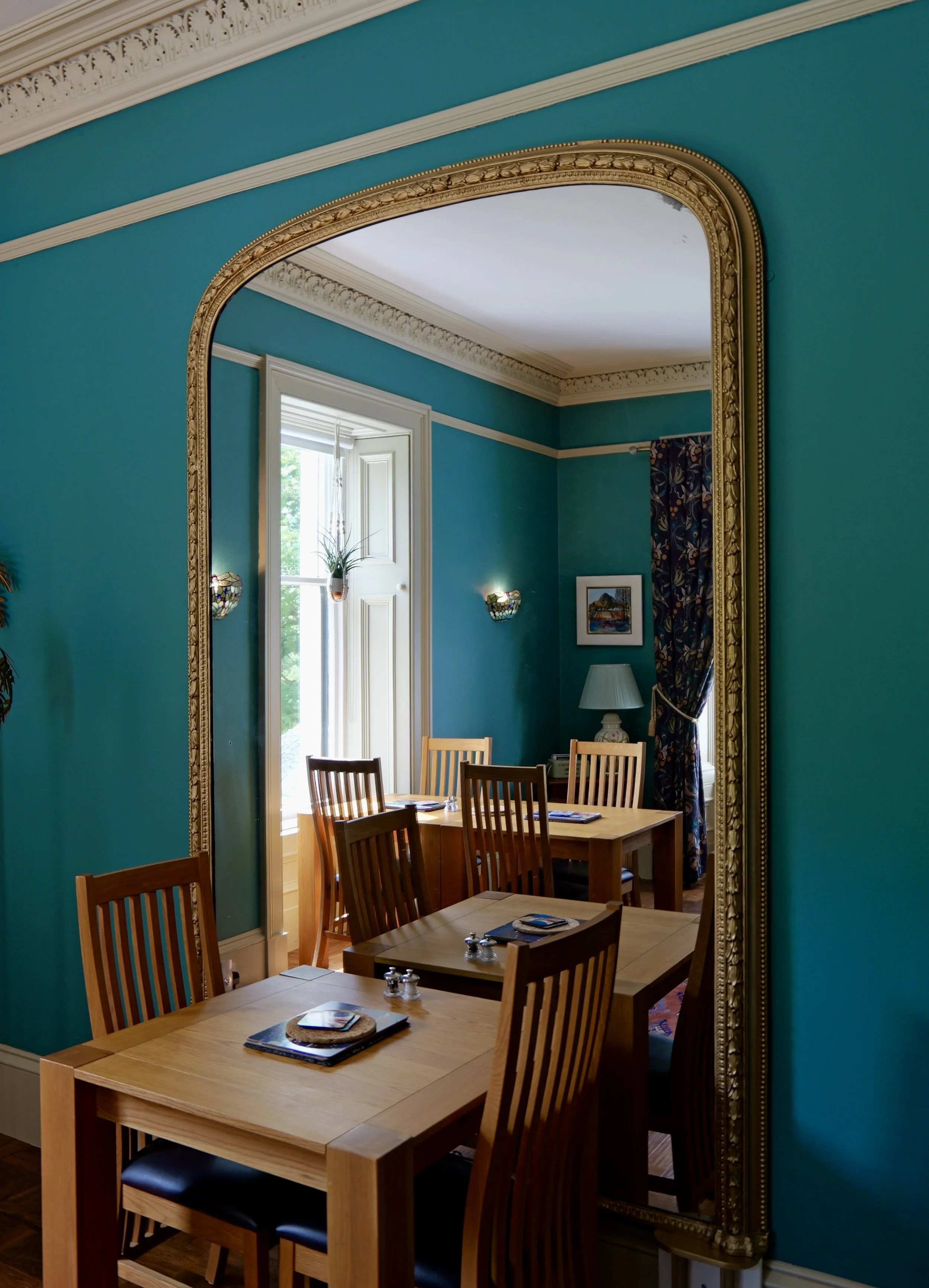 A large vintage mirror with ornate gold trim reflects a dining room with teal walls, a window with curtains, and a wooden table with chairs.