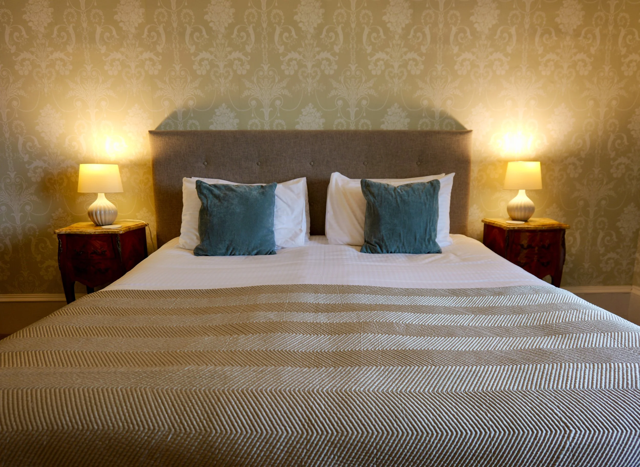 A neatly made bed with two teal pillows and white pillows, a beige textured blanket at the foot of the bed, a gray fabric headboard, and two matching table lamps on wooden nightstands.