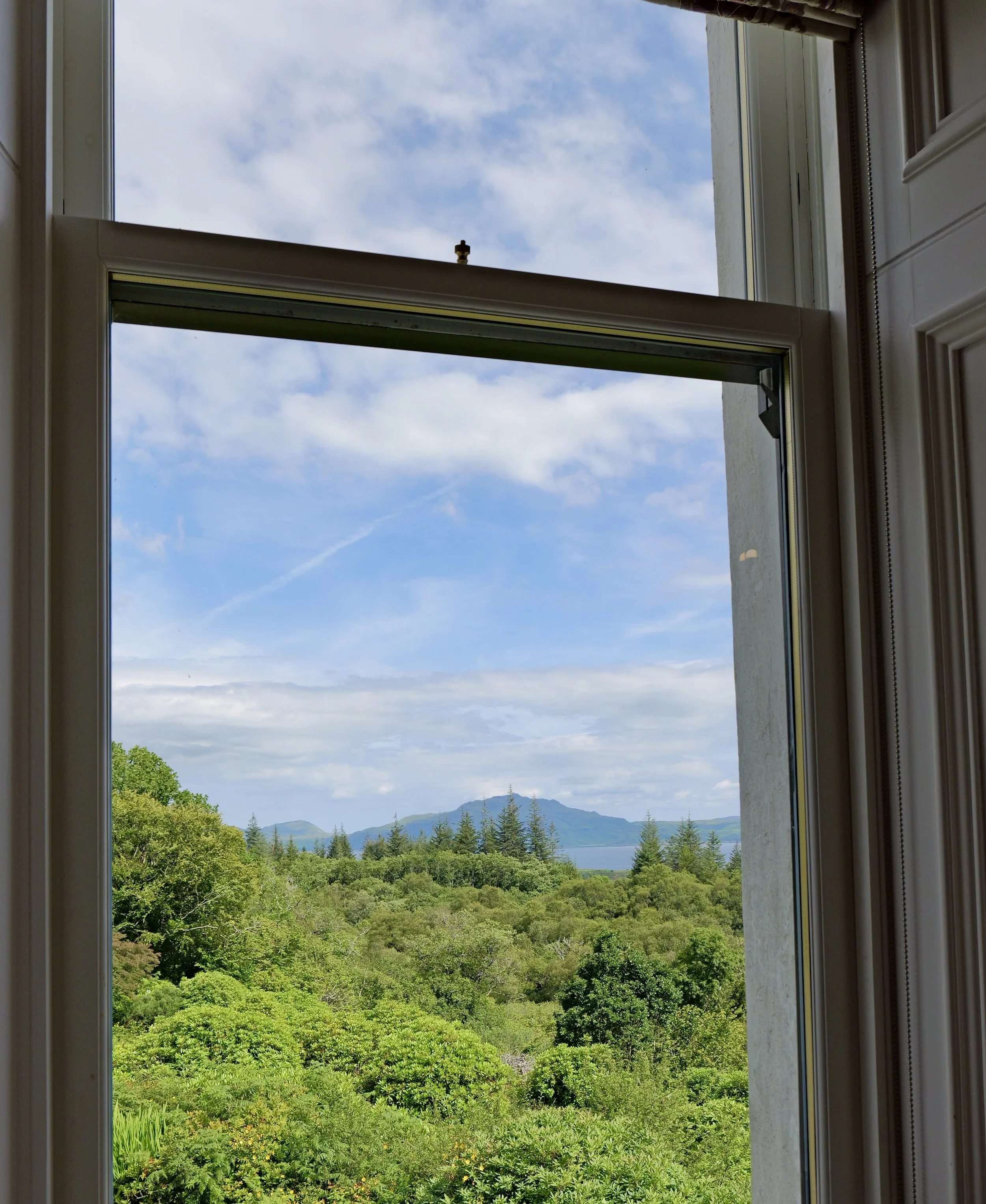 Open window showing green trees, distant mountains, and partly cloudy sky.