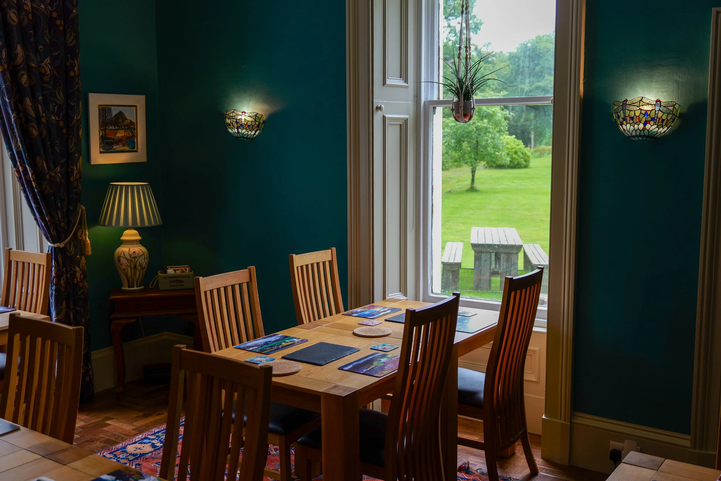A dining room with a wooden table set with game cards and cork coasters, surrounded by wooden chairs, and large window showing a lush green outdoor area with picnic table.