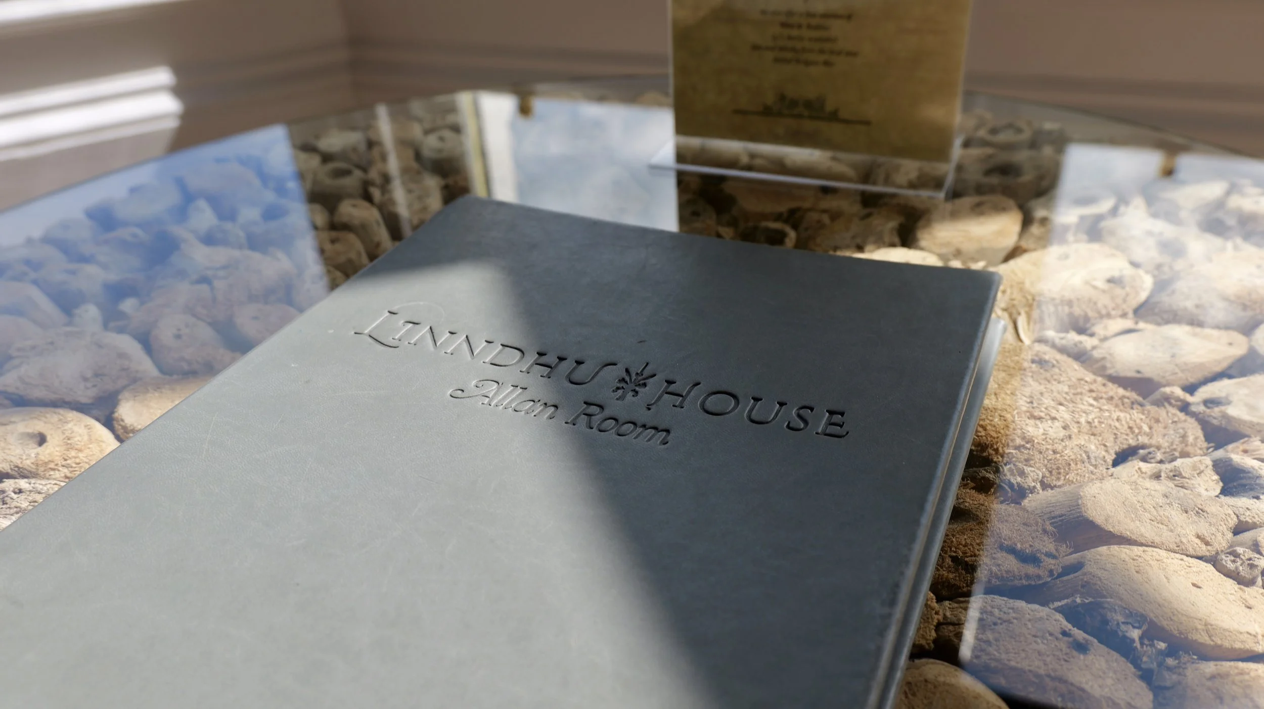 A guest book with the words 'LINDHU#HOUSE Alan Room' embossed on the cover, placed on a glass surface with rocks underneath and a sign in the background.