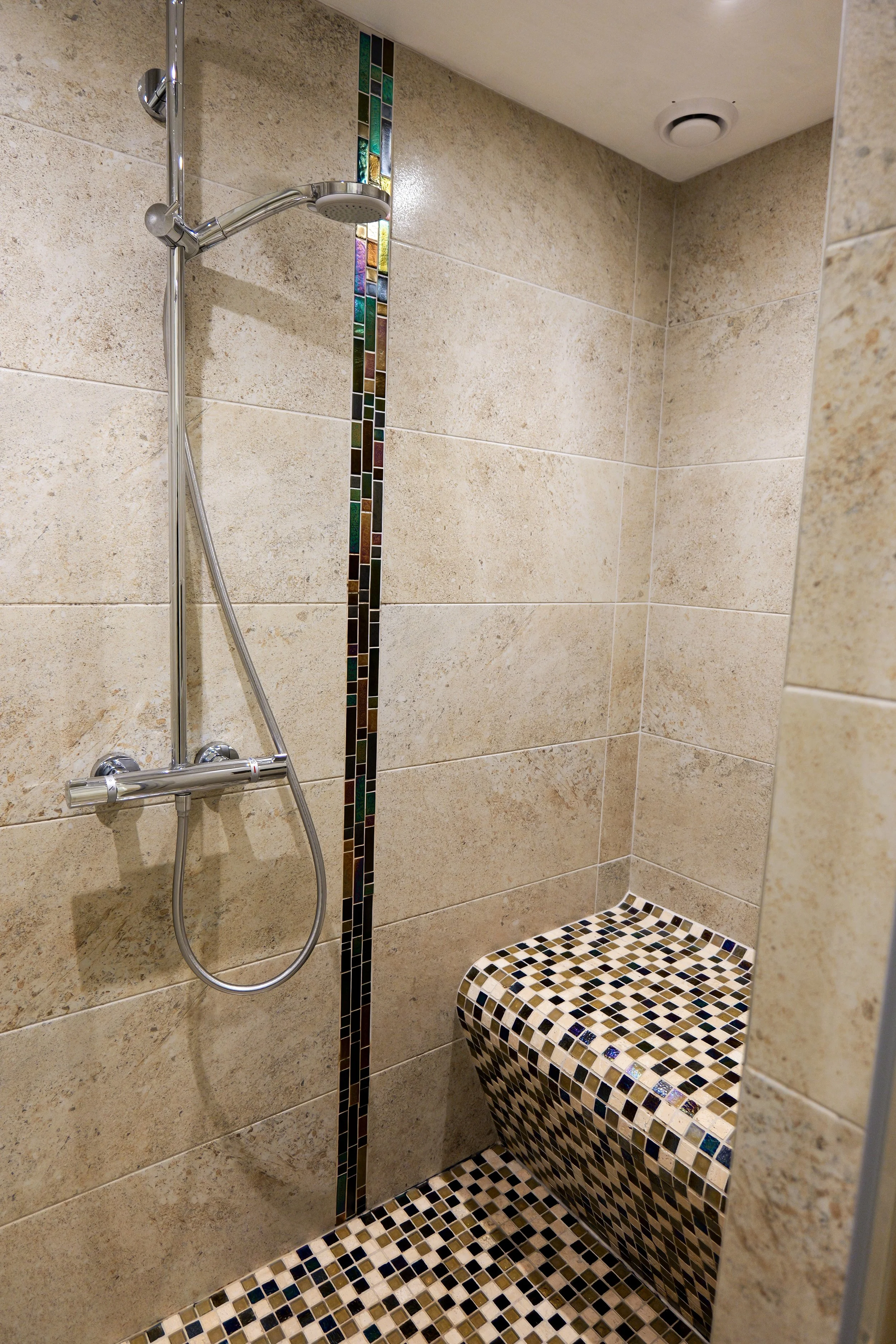 A corner shower with beige tiled walls, a vertical strip of colorful mosaic tiles, a handheld showerhead attached to a horizontal bar, and a seating ledge with a mosaic tile pattern.