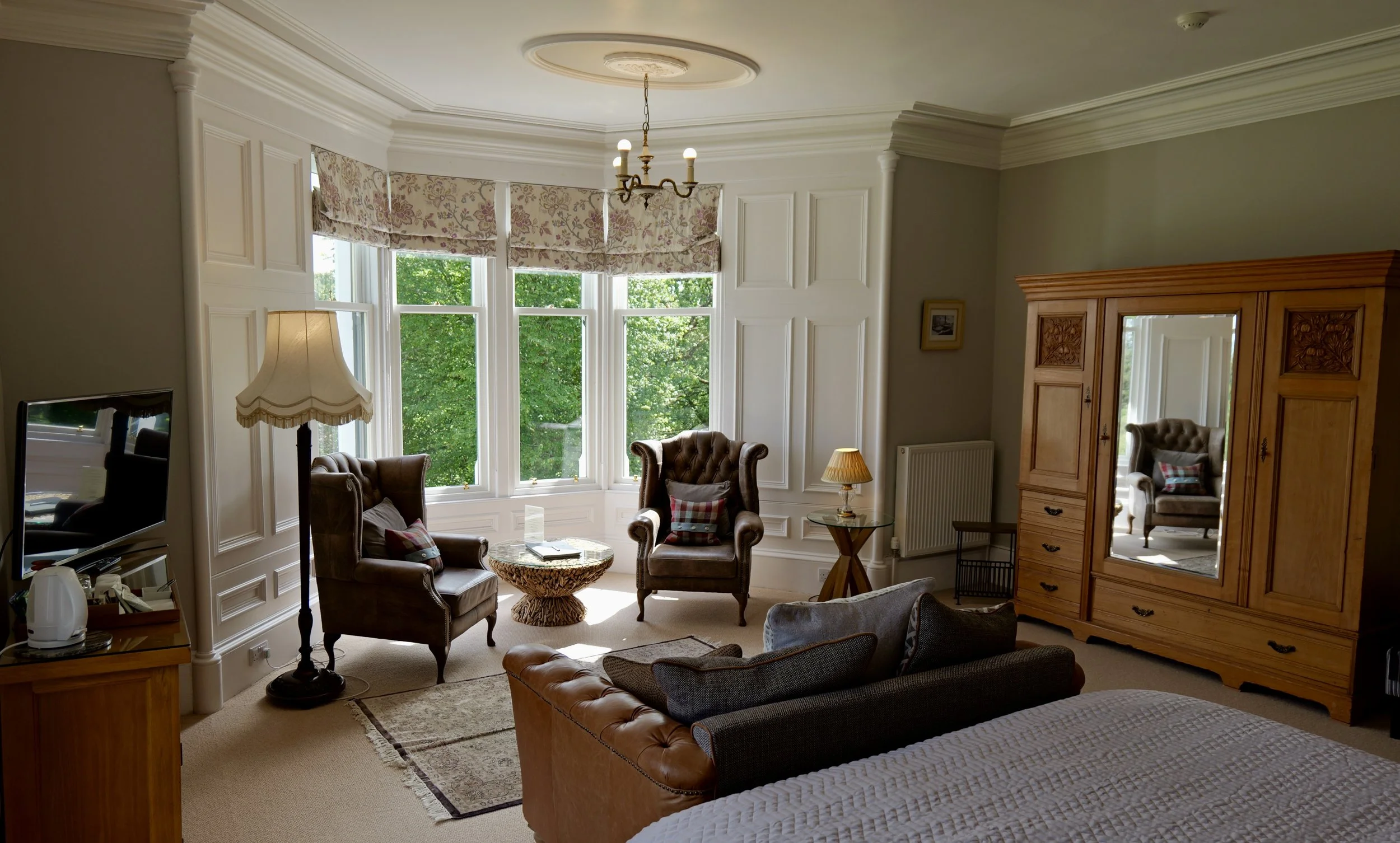 A cozy vintage-style living room with a large bay window, leather armchairs, a tufted sofa, wooden furniture, and a chandelier, with lush greenery outside.