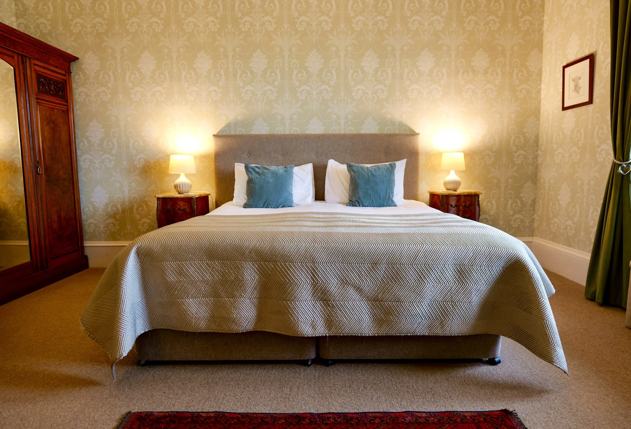 A neatly made bed with white sheets, four pillows (two with white pillowcases and two with teal pillowcases), and a beige bedspread. The bed has a gray upholstered headboard and is flanked by two wooden nightstands with white lamps. The room has beig