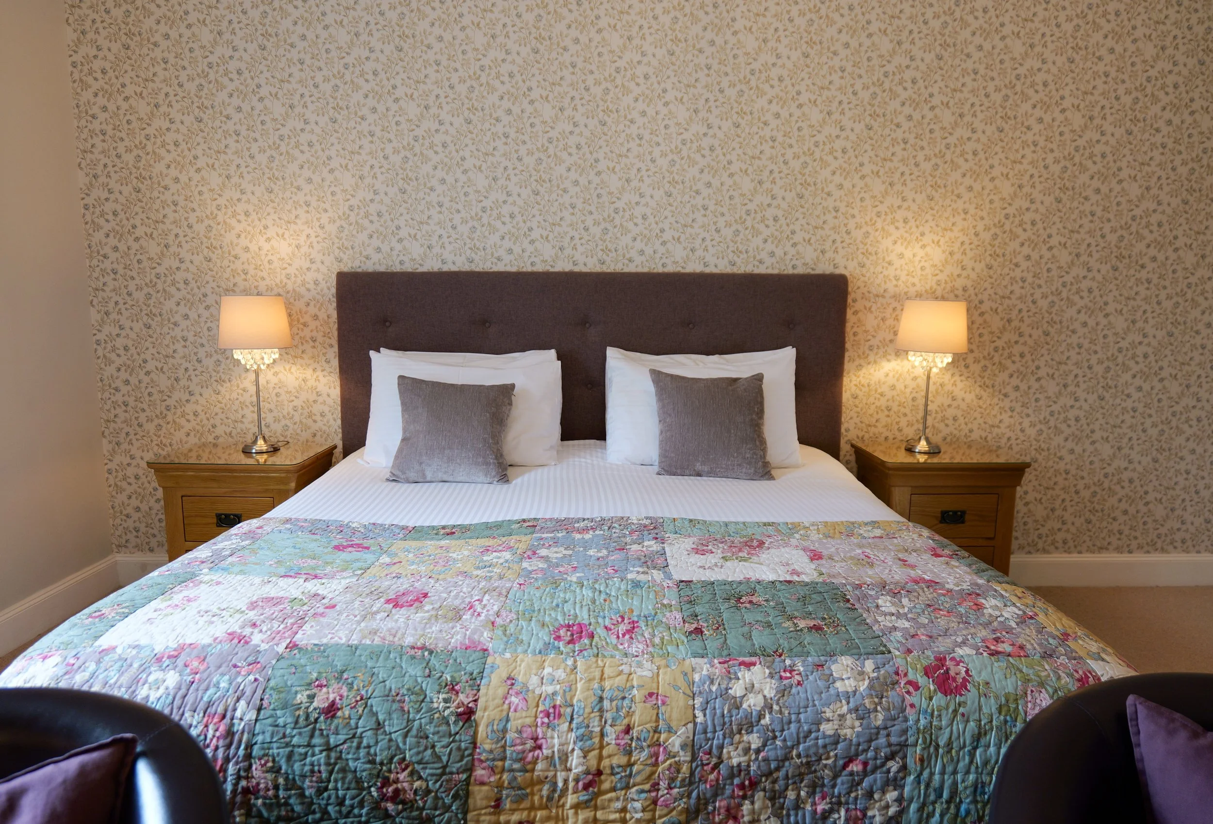 A cozy bedroom with a large bed featuring a colorful quilt, two white pillows with gray decorative pillows, a brown upholstered headboard, two wooden nightstands with lamps, and floral wallpaper.