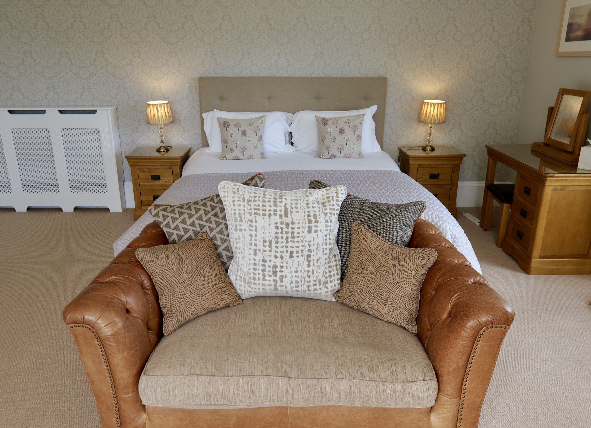 A bedroom with a bed, two bedside tables with lamps, a wooden desk with a mirror, and a brown leather sofa with decorative pillows facing the bed.