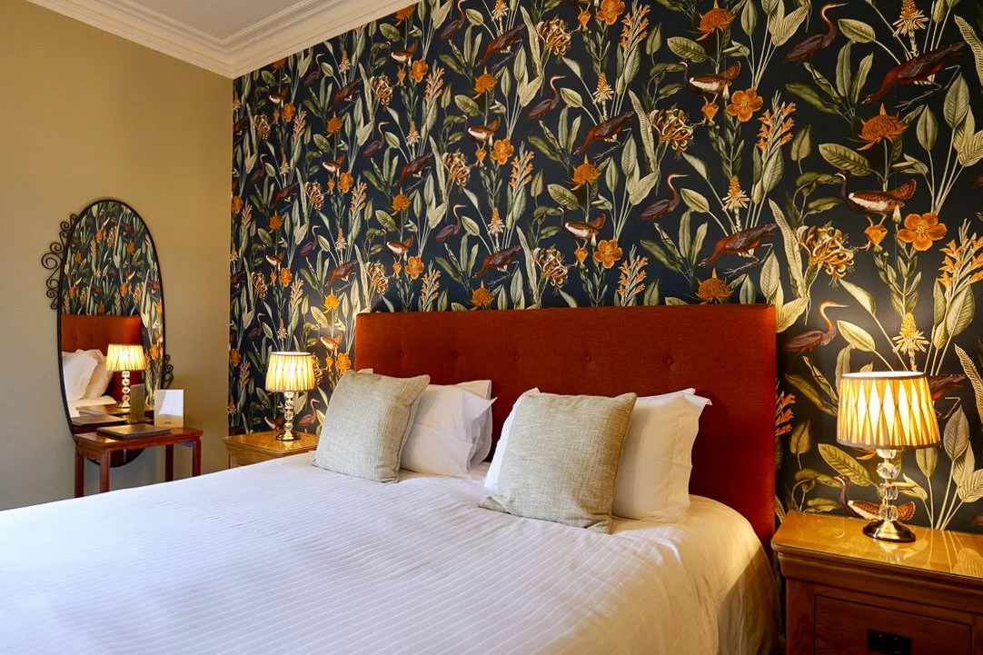 A bedroom with a large bed featuring a red headboard, floral wallpaper behind the bed, and two bedside tables with matching lamps. A mirror with a decorative frame is in the corner.