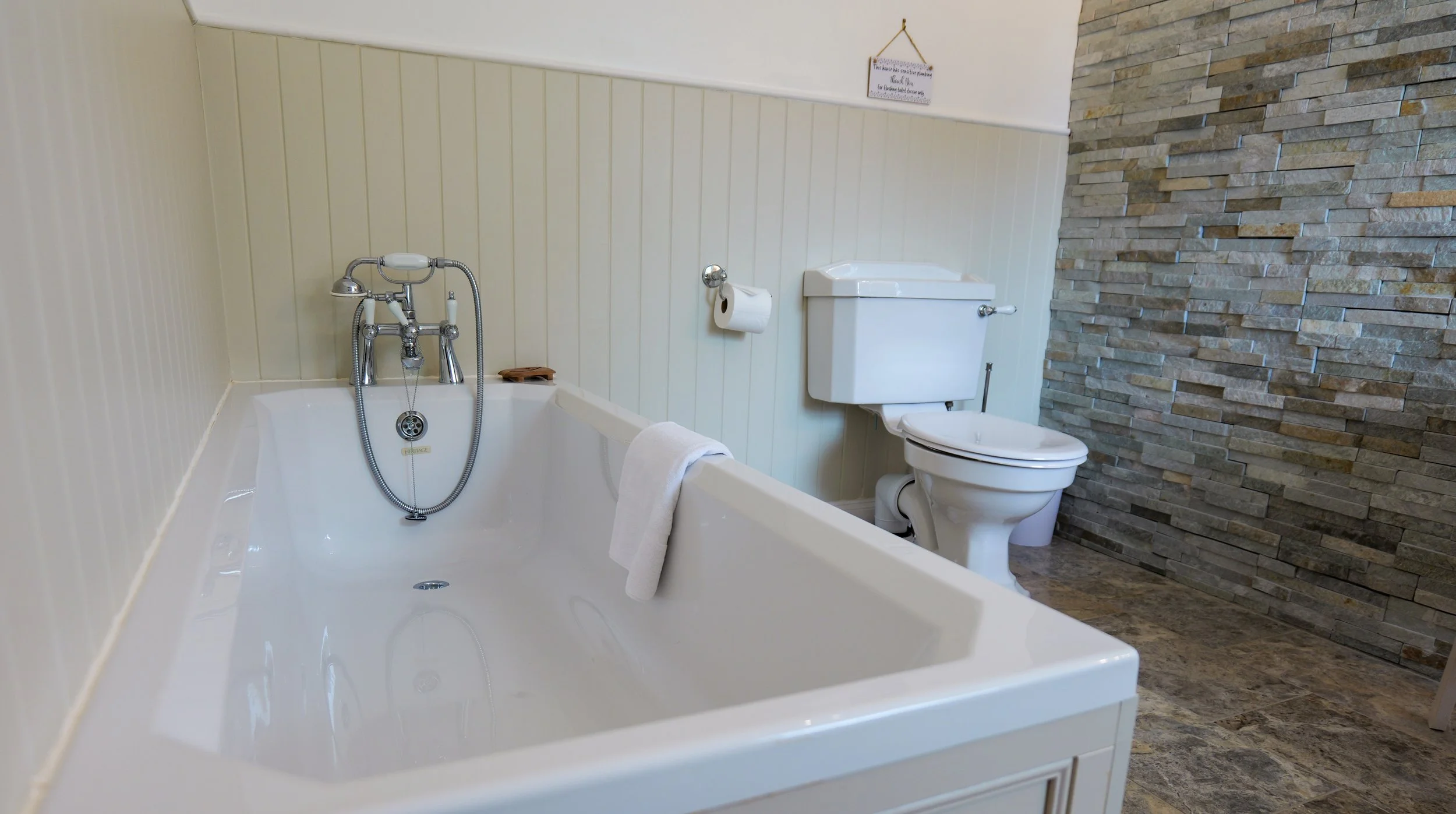 Bathroom with a white bathtub, a toilet, a wall with beige paneling, and a stone accent wall.