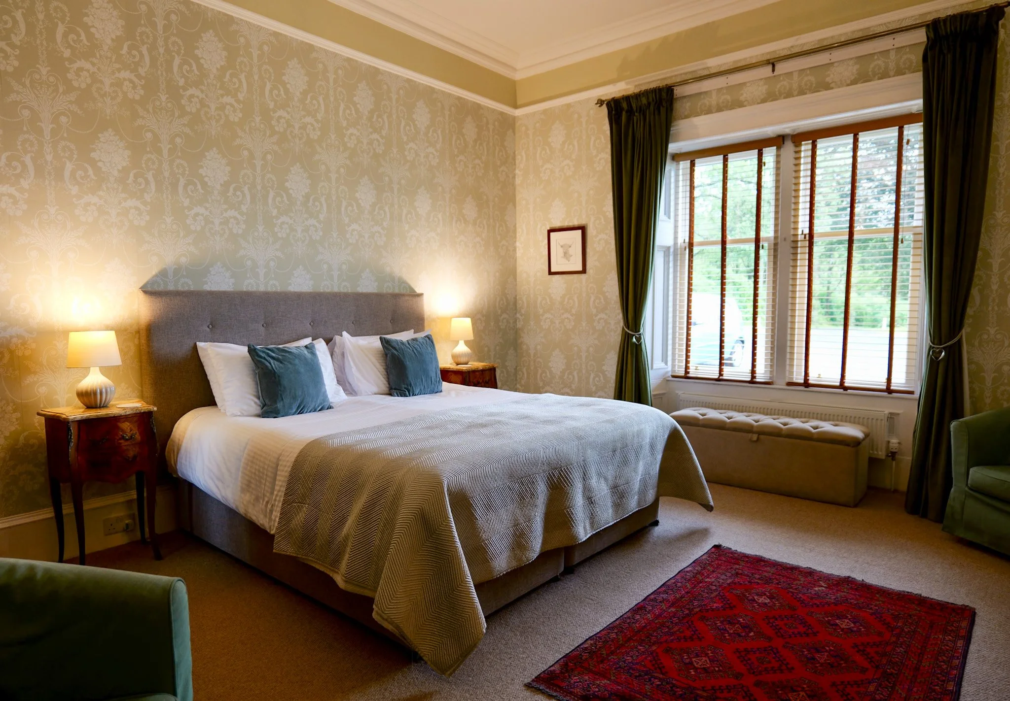 A cozy bedroom with a bed, pillows, and lamps, featuring wallpaper and a large window with green curtains and wooden blinds.