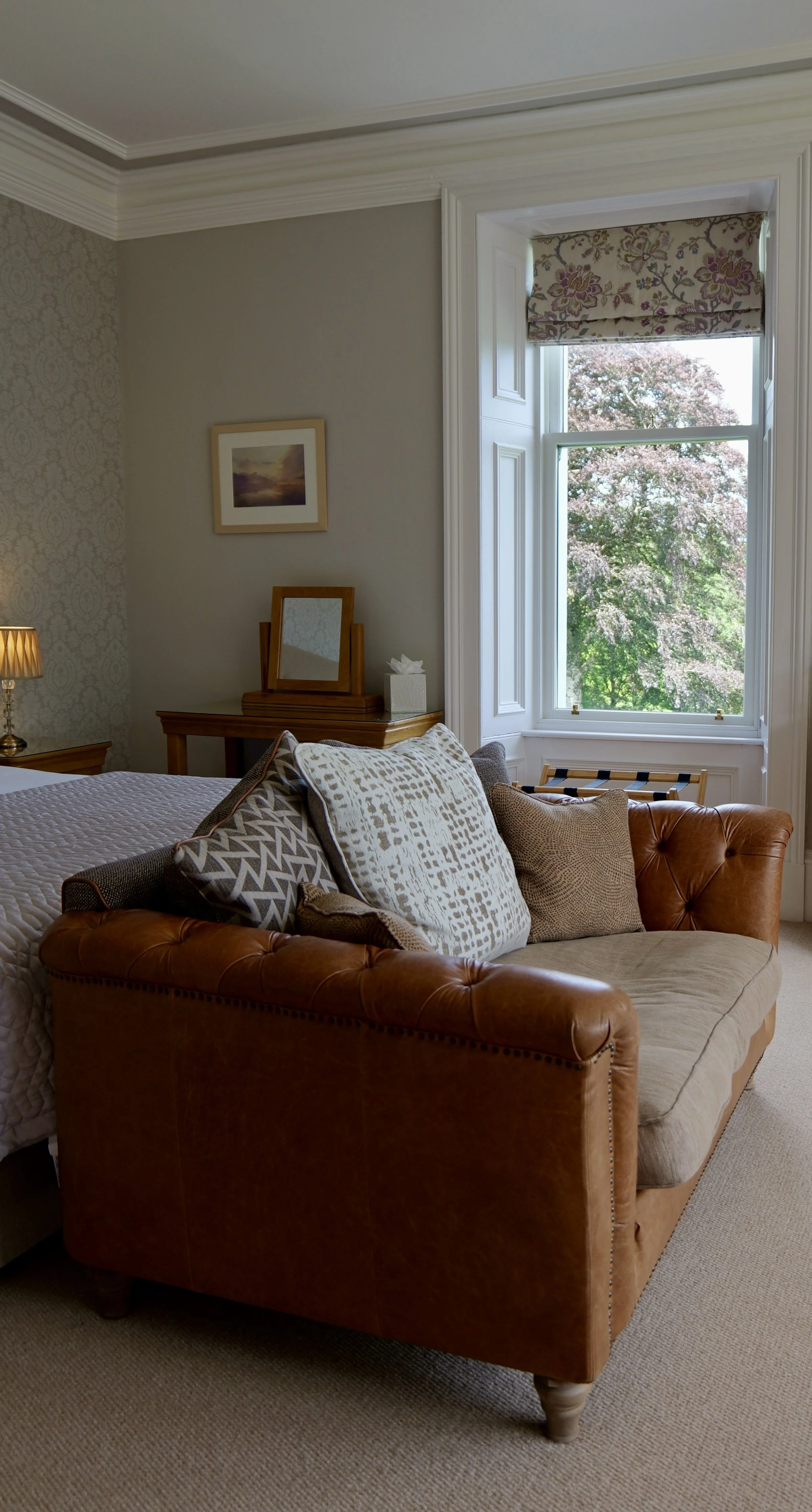 Living room with a brown leather sofa, patterned pillows, large window with floral valance showing trees outside, side table with lamp, and a bed partially visible.