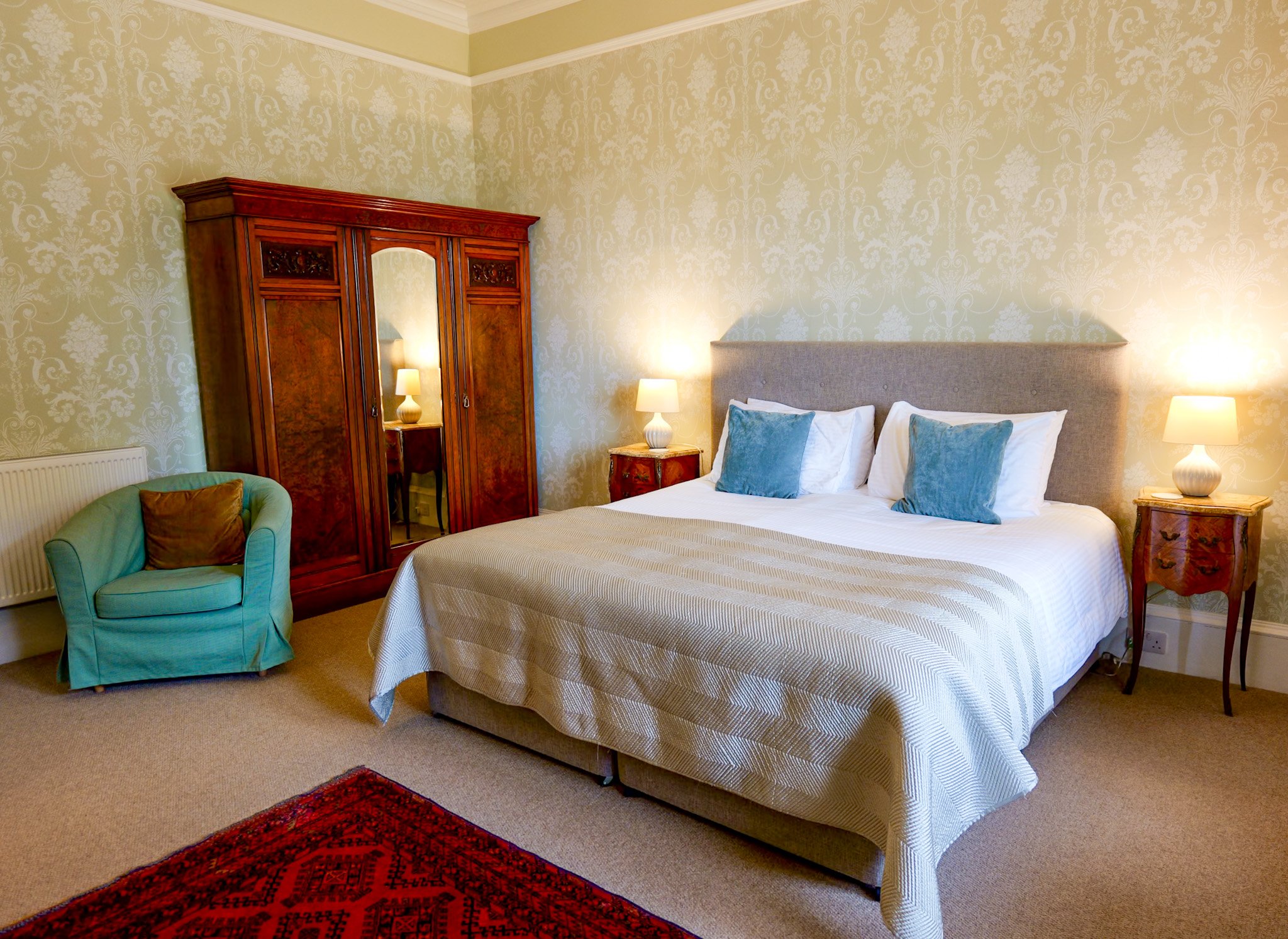 A well-made bed with a tufted gray headboard, white linens, and four blue pillows, flanked by wooden nightstands with lamps. There is an antique wooden wardrobe with a mirror reflecting a lamp, a green armchair with a brown pillow, a beige carpet, an