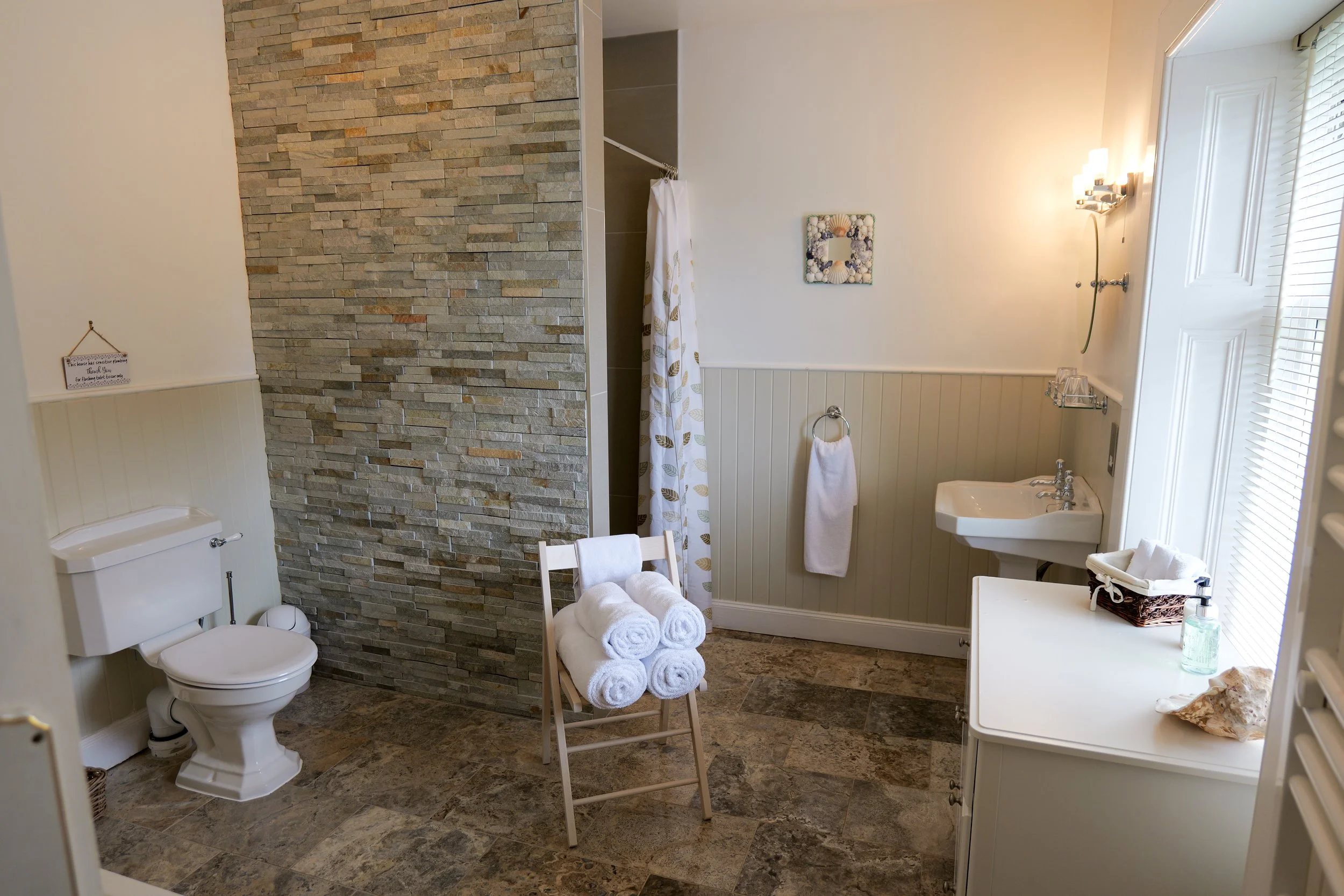 A bathroom with a toilet, a small sink, a towel rack, and a shower with a leaf-patterned curtain. There is a chair with rolled towels, a vanity with toiletries, a wall-mounted light, and a stone accent wall.