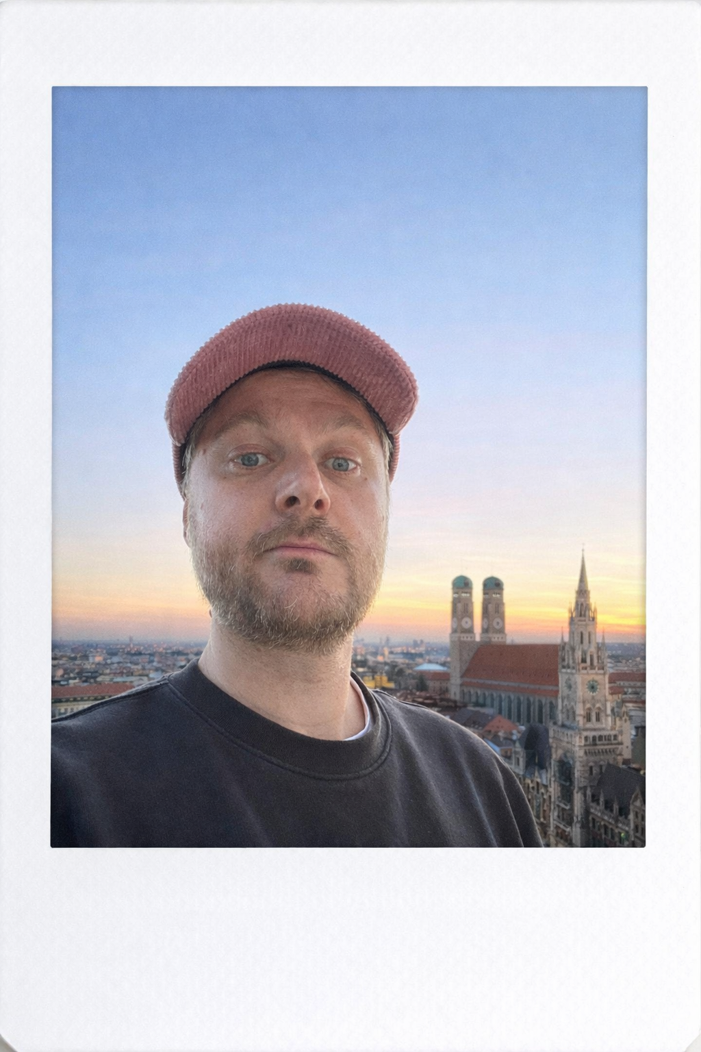 Ein Mann mit rosa Mütze und schwarzem sweatshirt steht auf einem Dach mit Blick auf eine Stadt bei Sonnenuntergang, im Hintergrund sind zwei Kirchtürme und ein historisches Gebäude sichtbar.