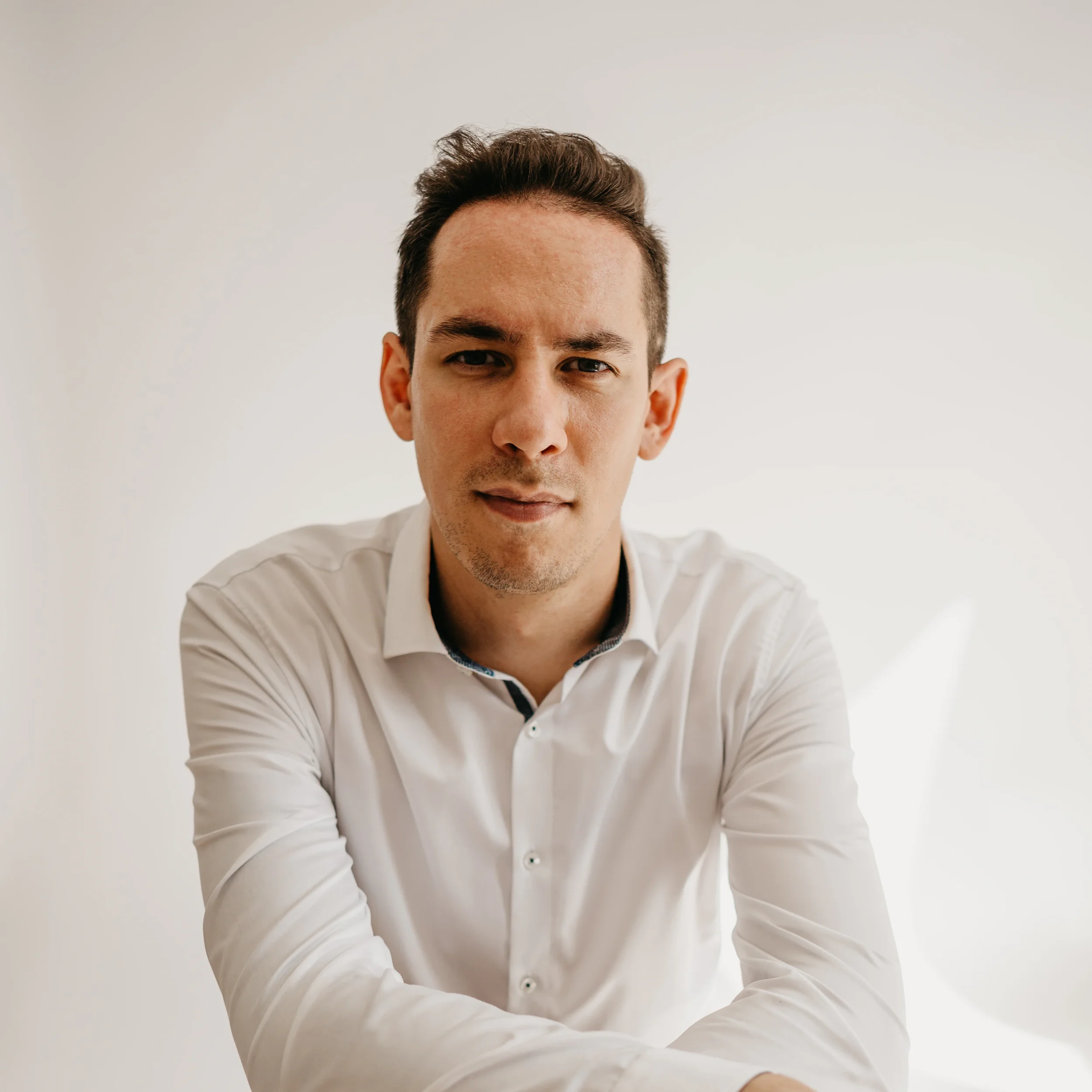Professional coach looks directly at the camera, sitting in front of a solid white background. He wears a white dress shirt.