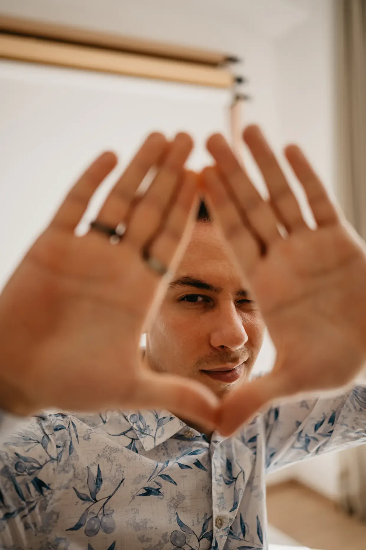 A young man with light skin and dark hair wearing a white shirt with a blue floral pattern is looking through a heart shape made by his hands while smiling and winking.