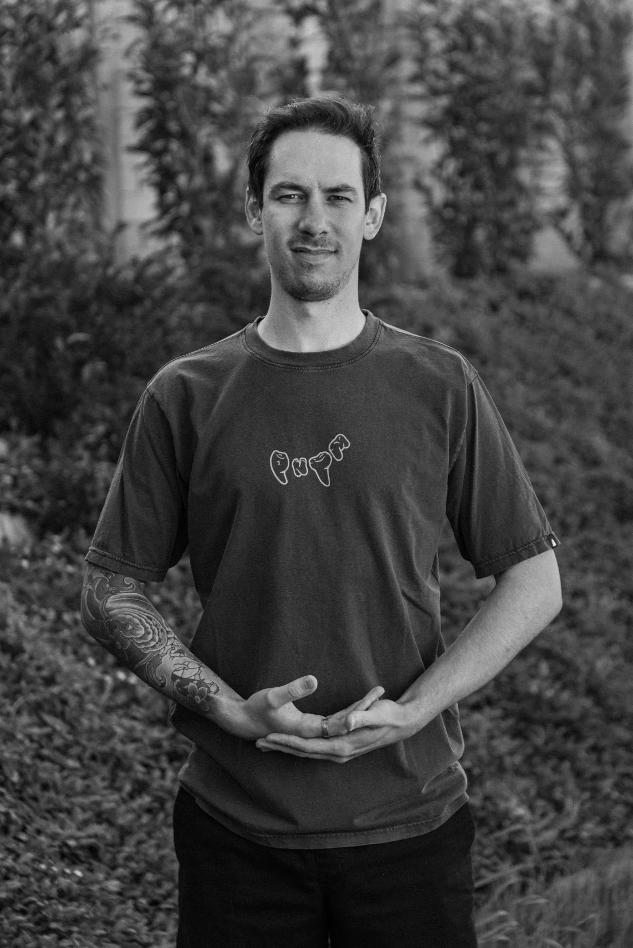 Professional coaching looking at directly at the camera. He holds his arms in a cradle position signaling a holding gesture. The picture is black and white, the background is a wall of plants.