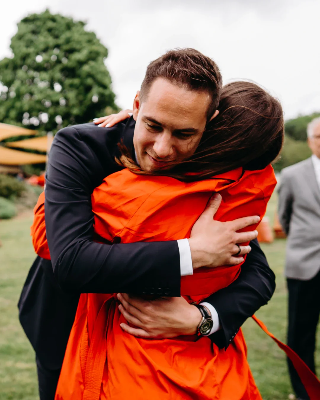 Professional coach in deep blue suit hugs his friend warmly who is in a bright orange coat.