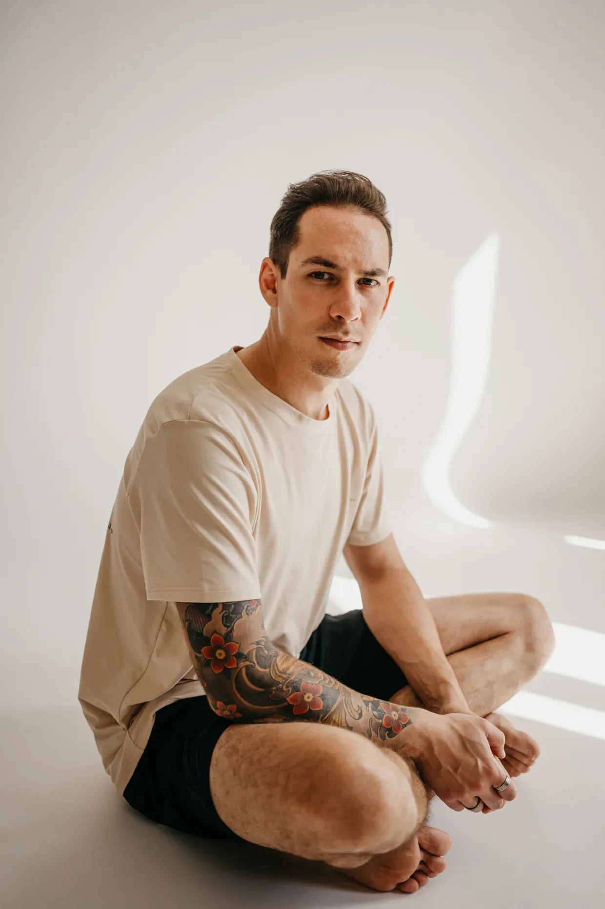 A young man with a full-sleeve oriental tattoo on his right arm sitting cross-legged on the floor in front of a plain light background.