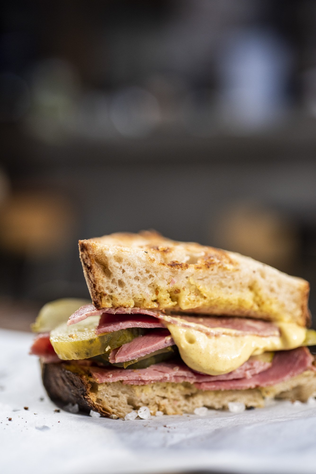 Close-up of a sandwich with ham, pickles, cheese, and toasted bread on parchment paper.