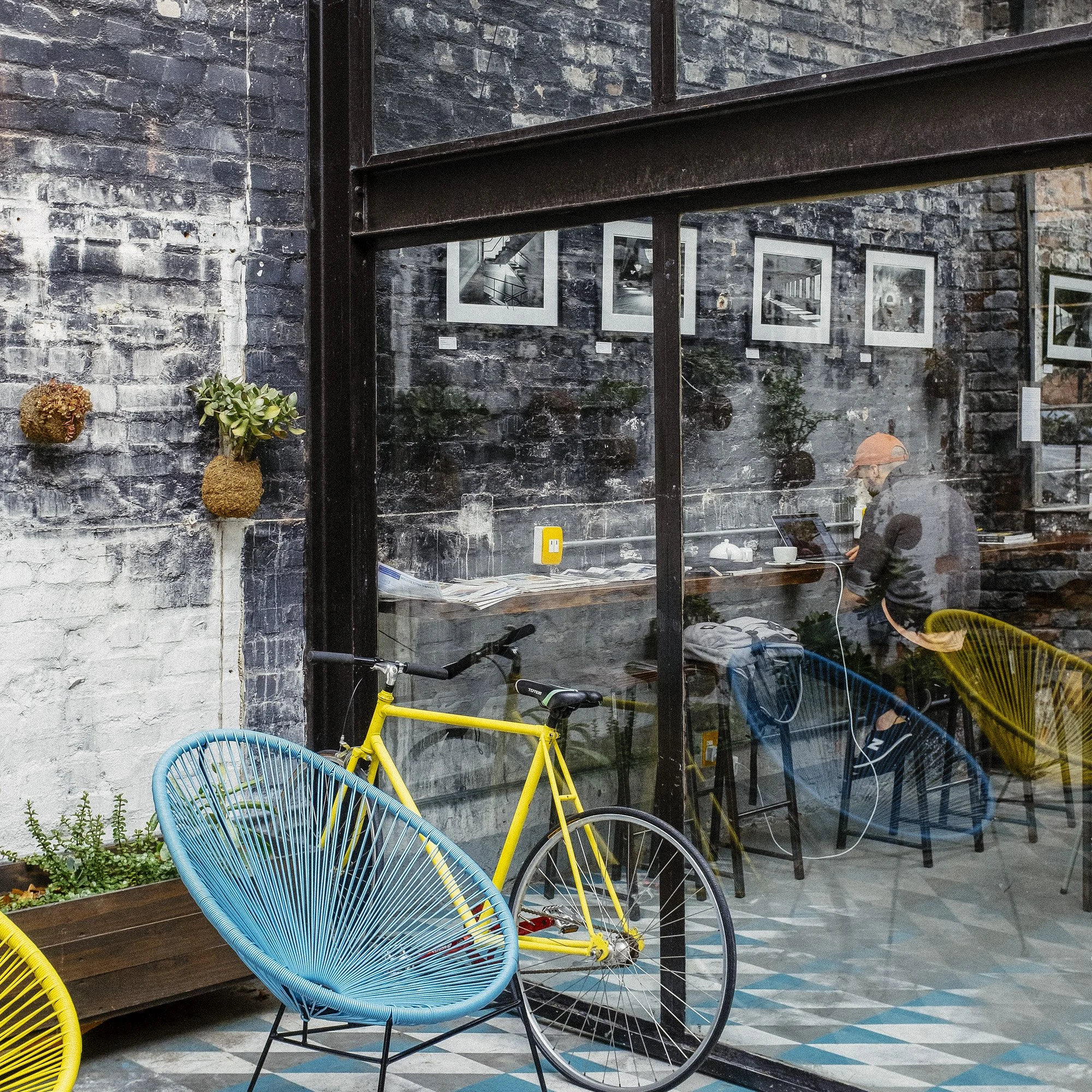 View through a glass window showing a woman sitting at a table inside a cafe, working on a laptop. Outside, a yellow bicycle is parked next to a vibrant blue outdoor chair, against a studio-style brick wall with wall-mounted plants and framed black-a