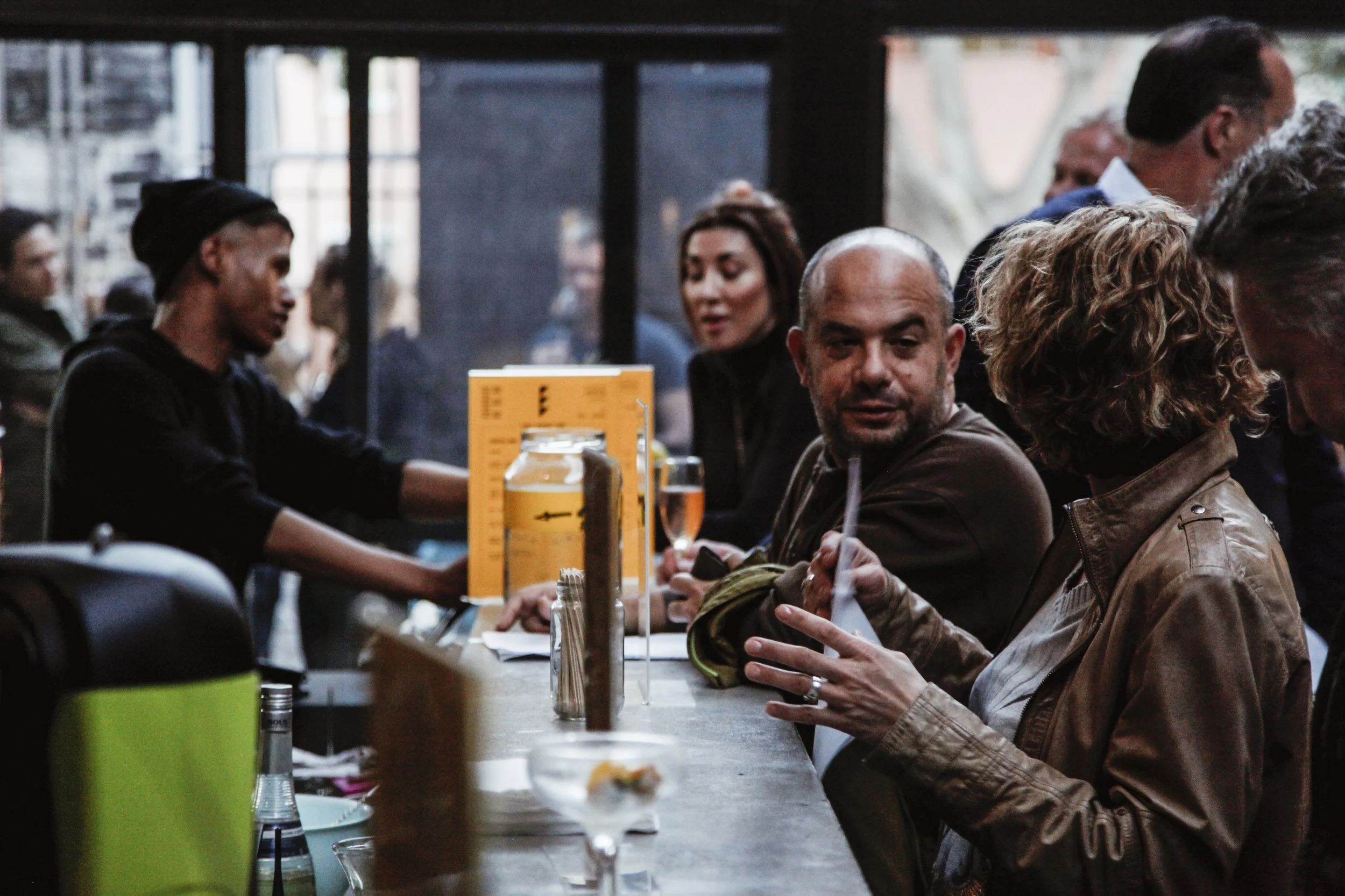 People sitting at a bar or restaurant counter, some talking and one woman using a tablet. Indoor setting with drinks and condiments on the counter.