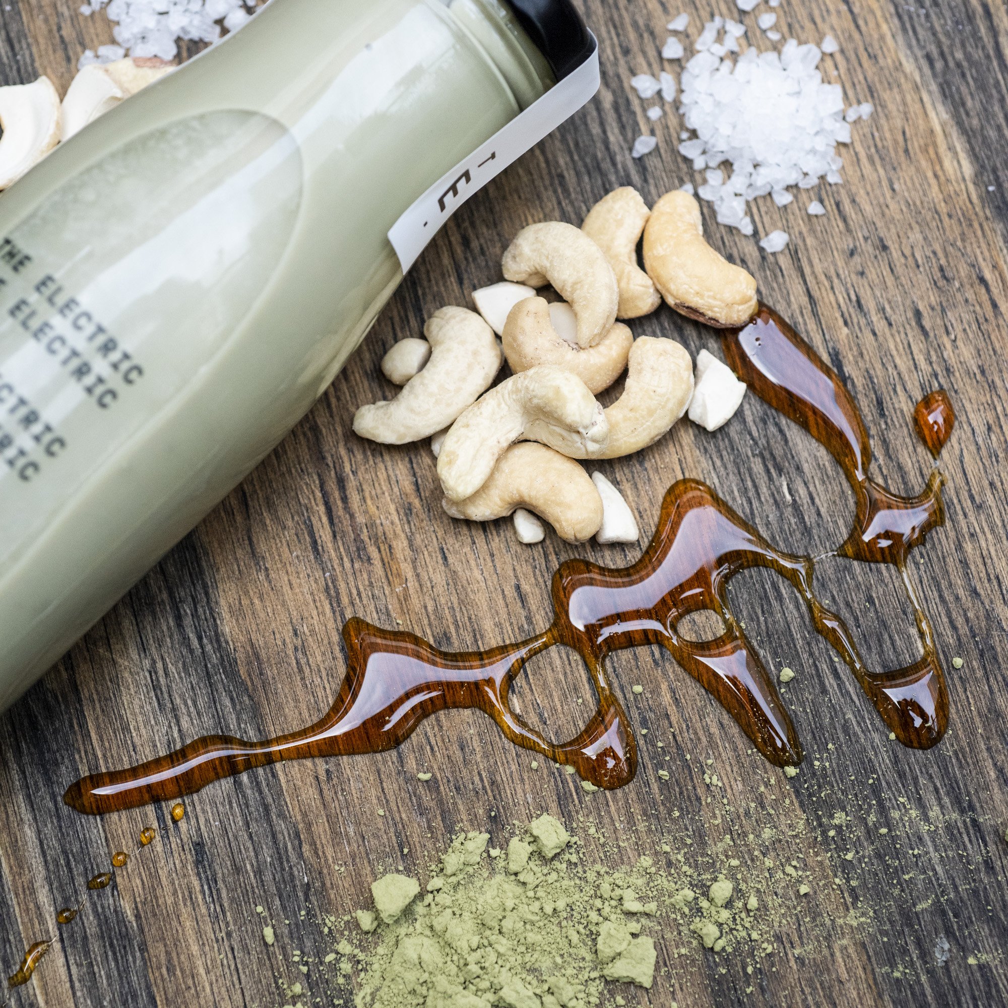 Assorted nuts, sea salt, honey, and ground matcha powder scattered on a wooden surface with a bottle of green juice