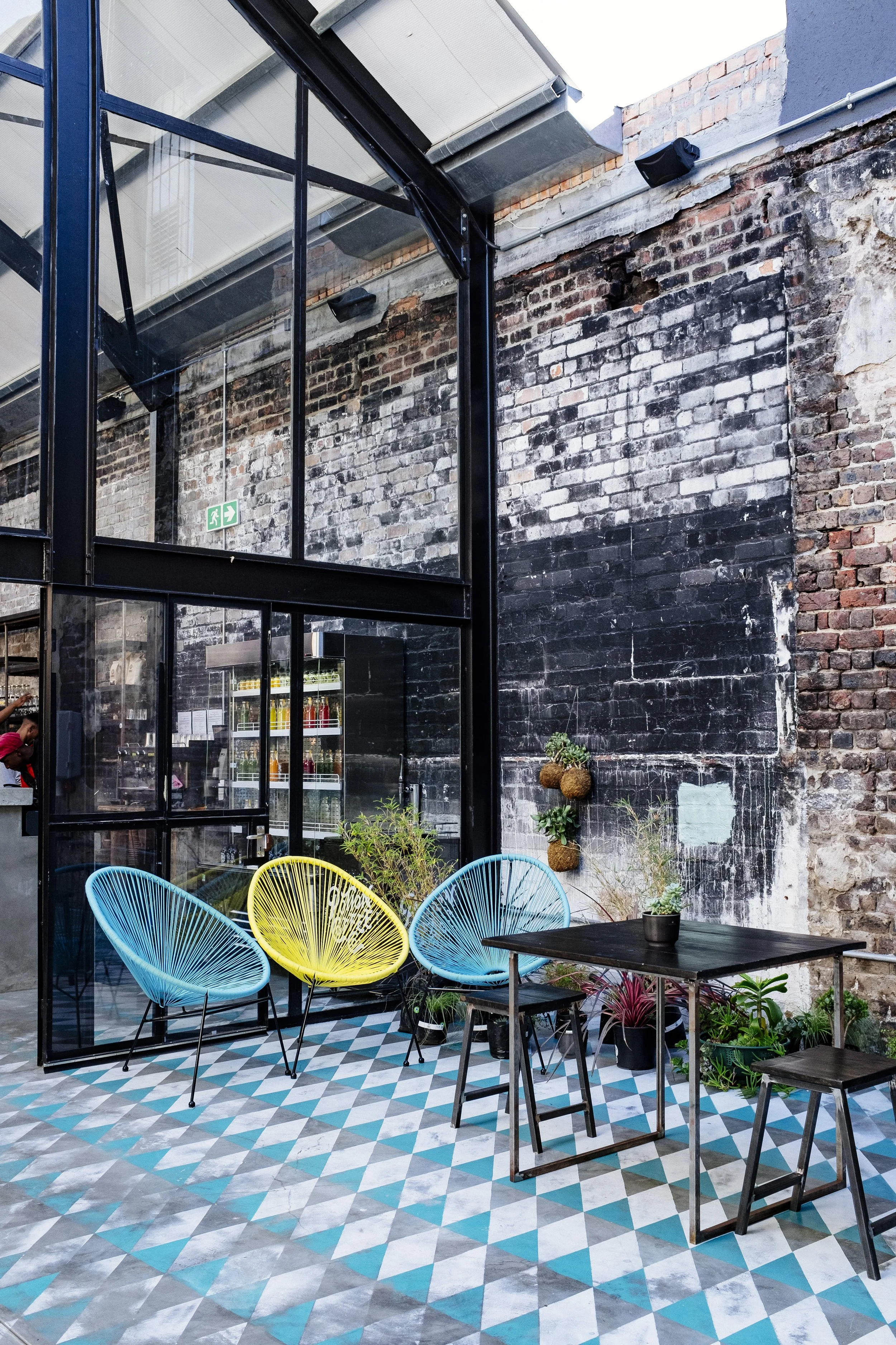 Indoor patio area with a black metal and glass partition, colorful chairs, and potted plants, against a weathered brick wall with a mural and hanging plant pots.