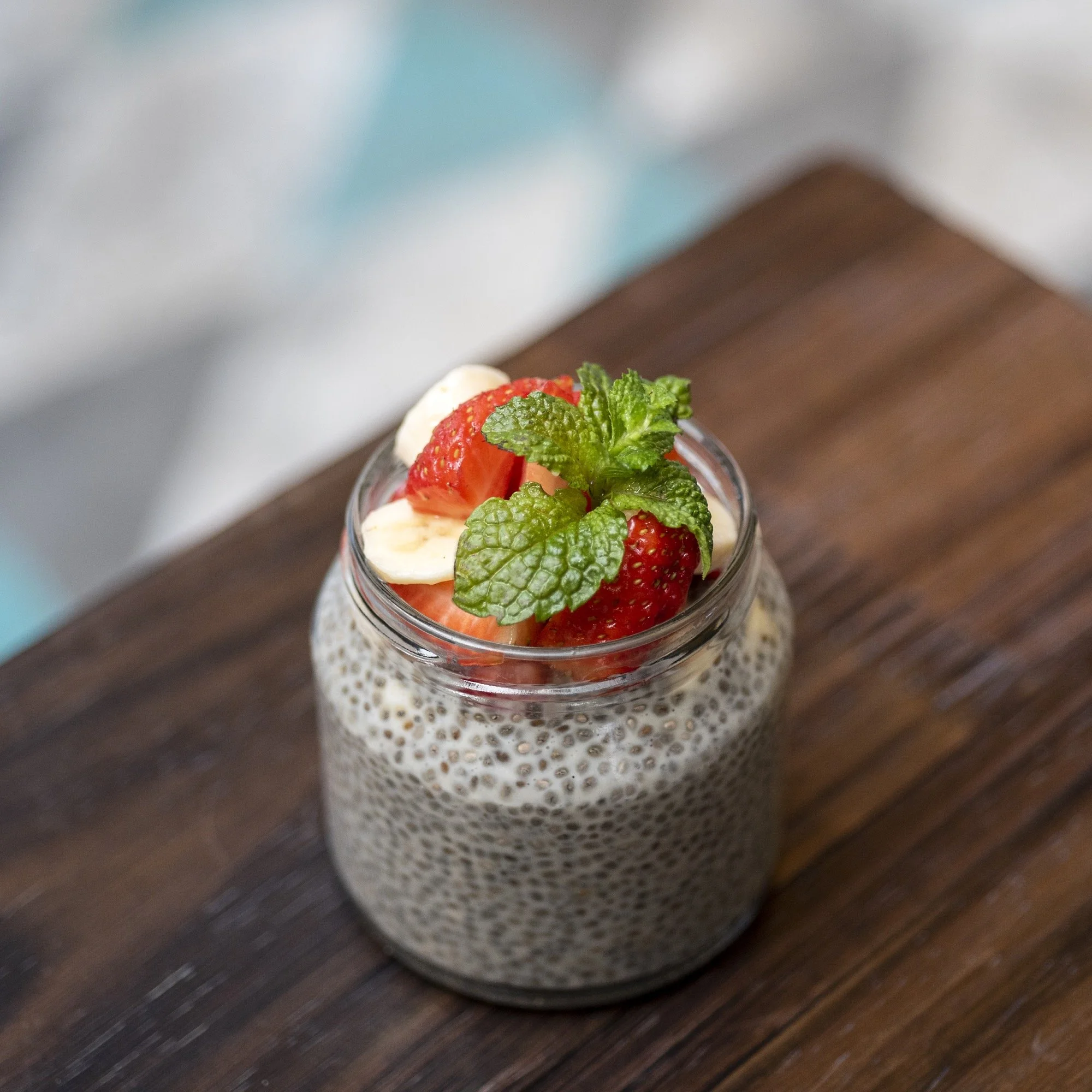 Chia seed pudding topped with strawberries, banana slices, and a sprig of mint in a glass jar on a wooden surface.