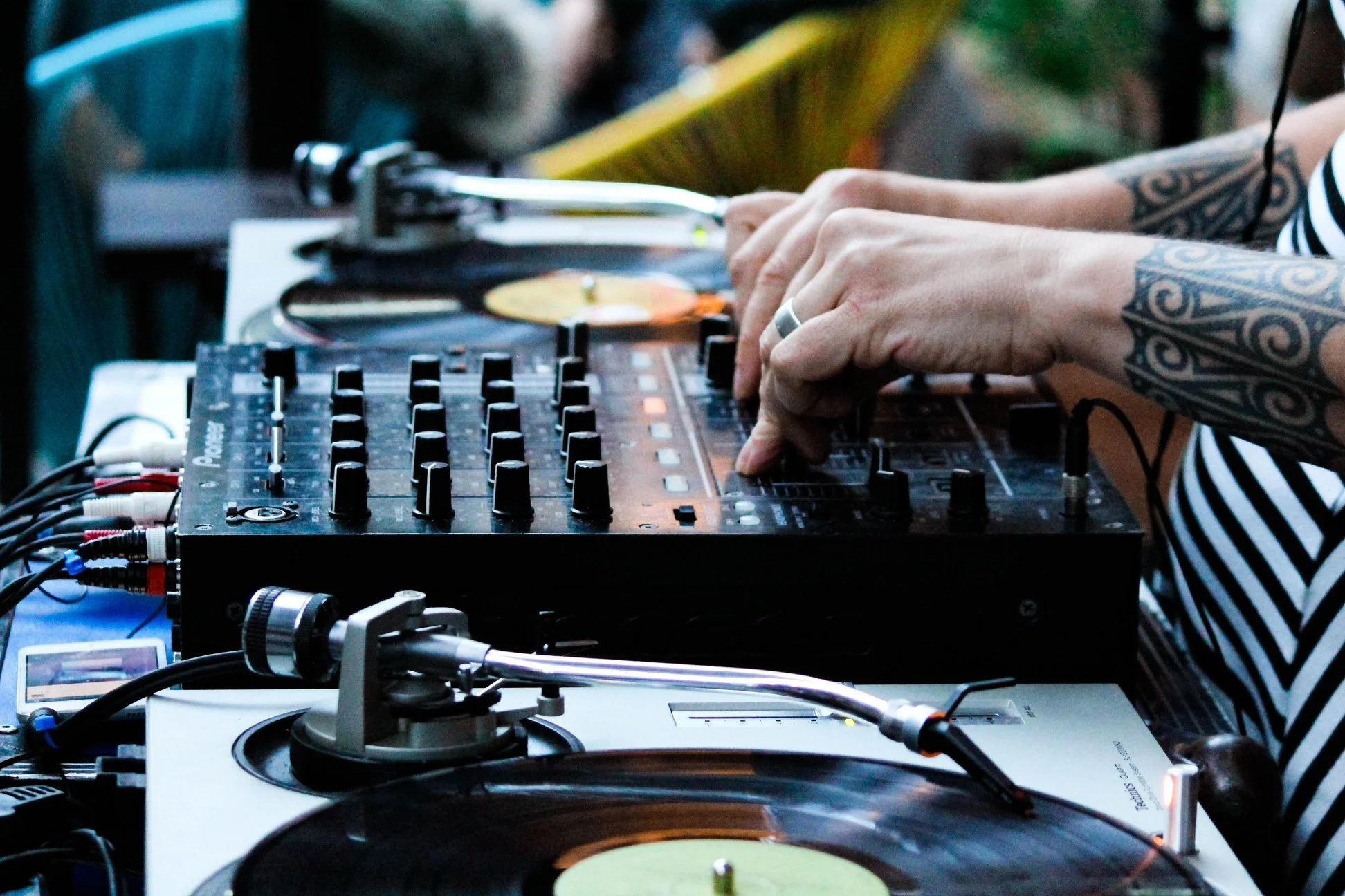 Close-up of a DJ mixing music with a turntable and a DJ controller, with a person adjusting the controls, wearing a striped shirt and tattooed arms.