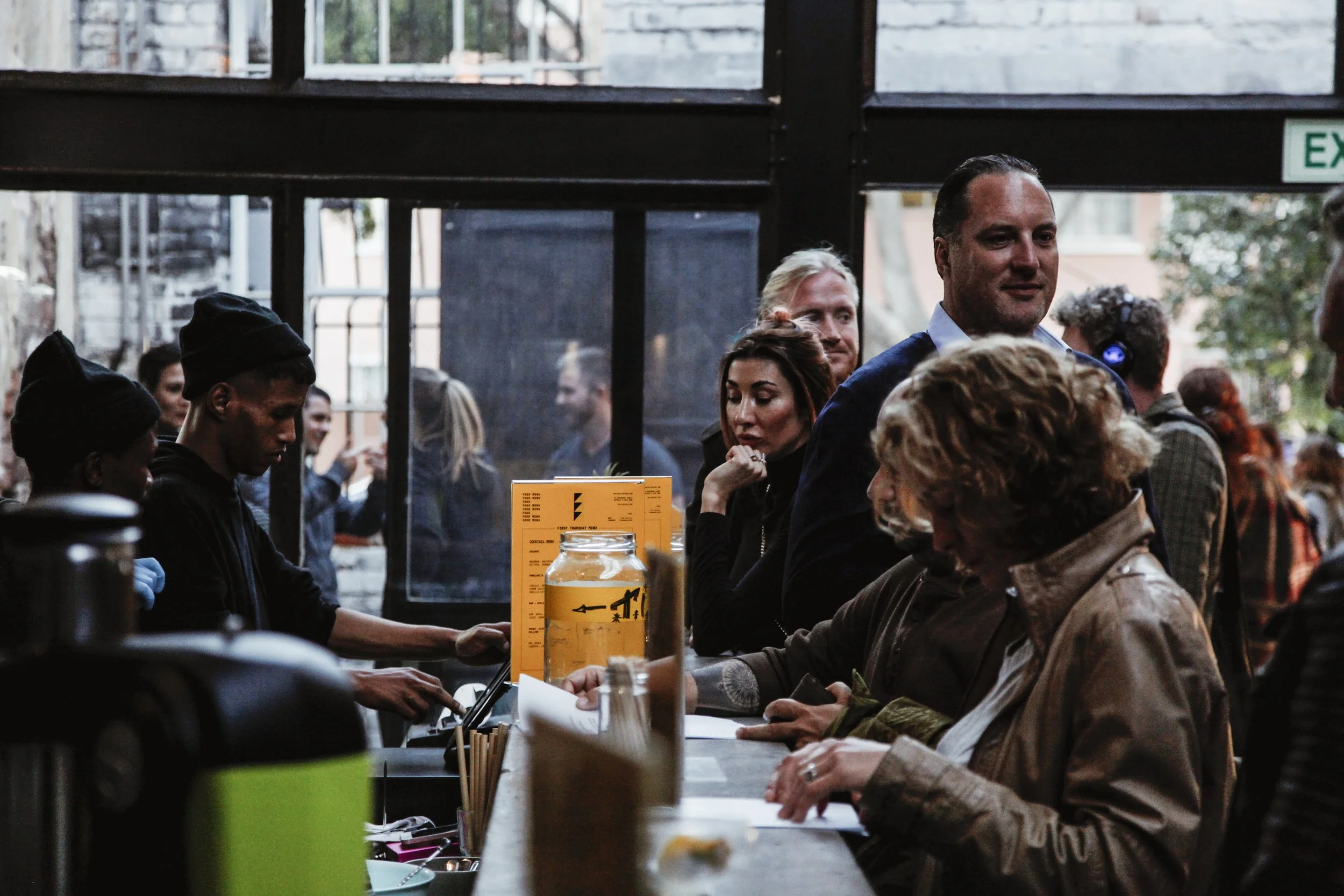 People standing in line at a cafe or restaurant counter inside a dimly lit venue, with a glass jar, yellow paper, and utensils on the counter.