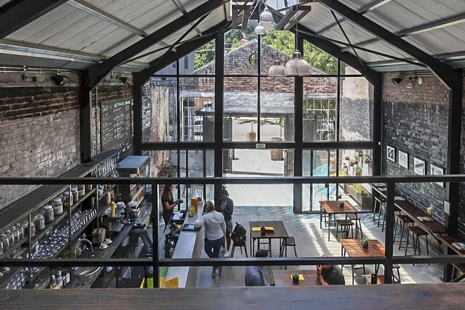 Interior of a modern coffee shop with high ceilings, brick walls, large windows, and a few customers. A barista is preparing drinks behind the counter, and tables are sparsely arranged with small potted plants. Natural light floods in through the windows.