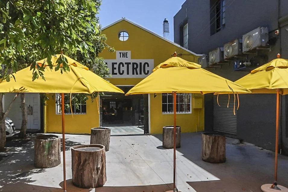 Yellow building with large sign reading "The Electric," three yellow umbrellas, wooden stumps as tables, and trees providing shade.