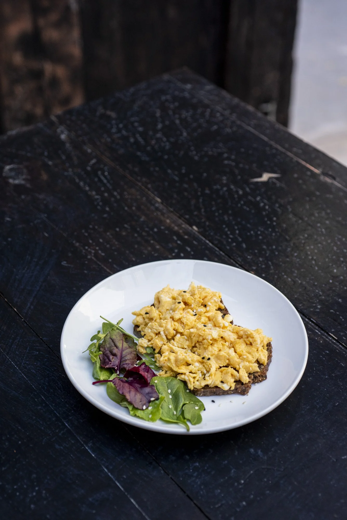 Scrambled eggs on toast with a side of mixed greens on a white plate, on a dark wooden table.