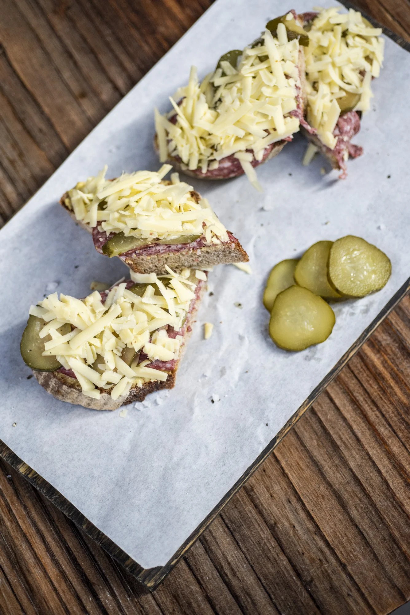 Open-faced sandwiches topped with shredded cheese, pickles on the side, served on a wooden tray lined with parchment paper.